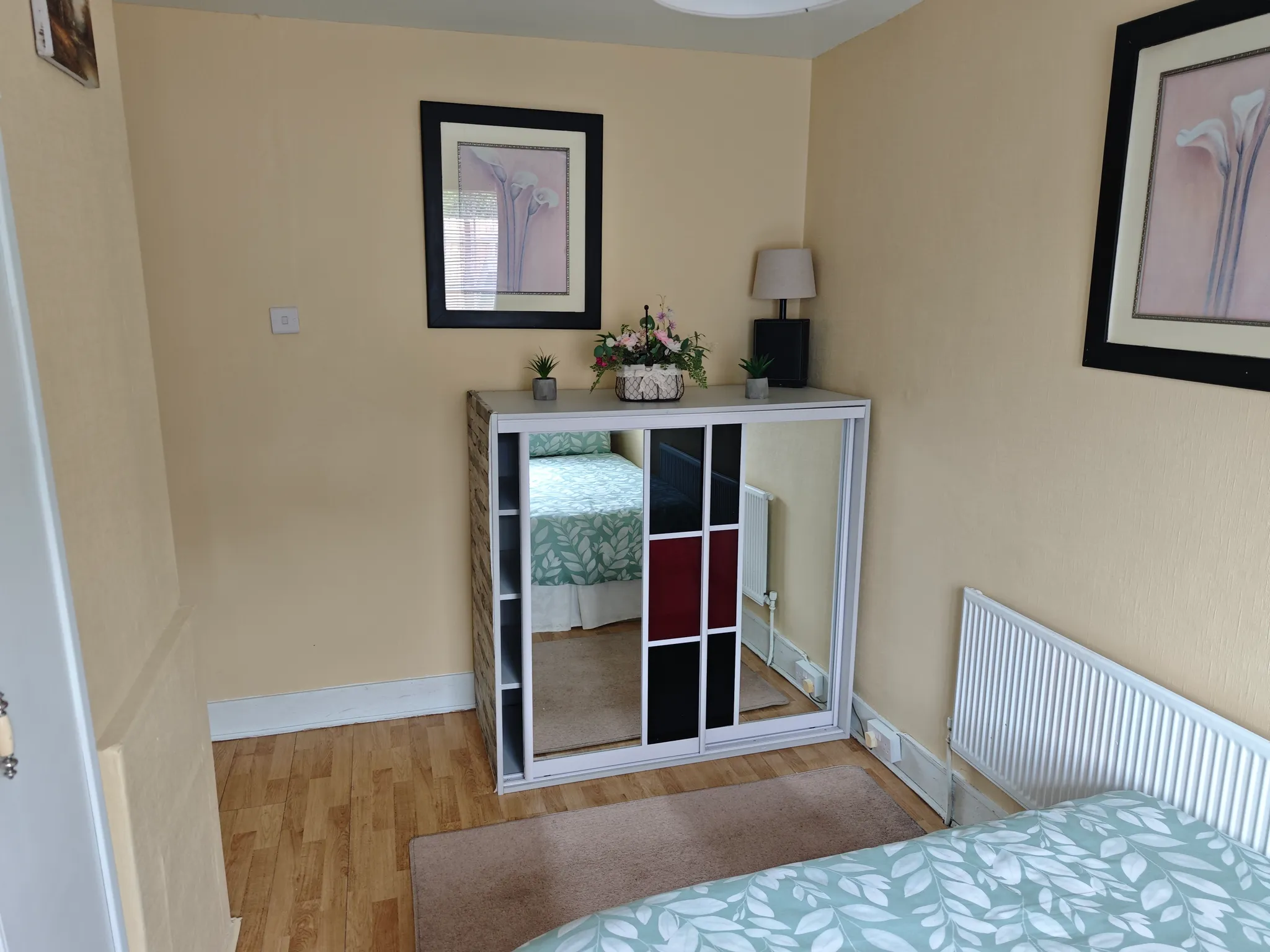 Bedroom corner with mirrored sliding wardrobe, framed wall art, a small lamp, a radiator and part of a bed with a leaf-pattern duvet.