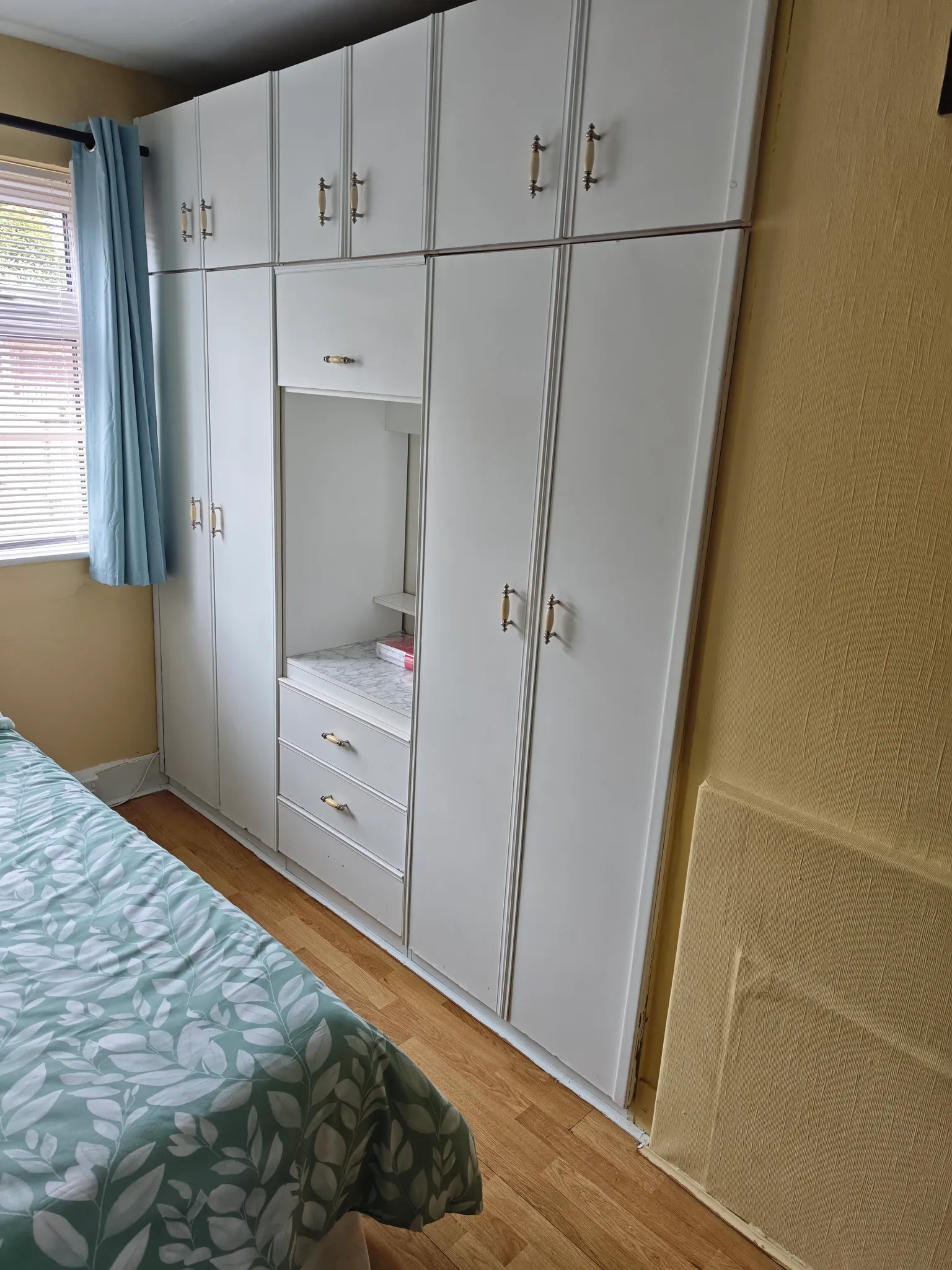 Bedroom with built-in white wardrobes and drawers, partial view of a bed with green patterned cover, and a window with blinds and a blue curtain.