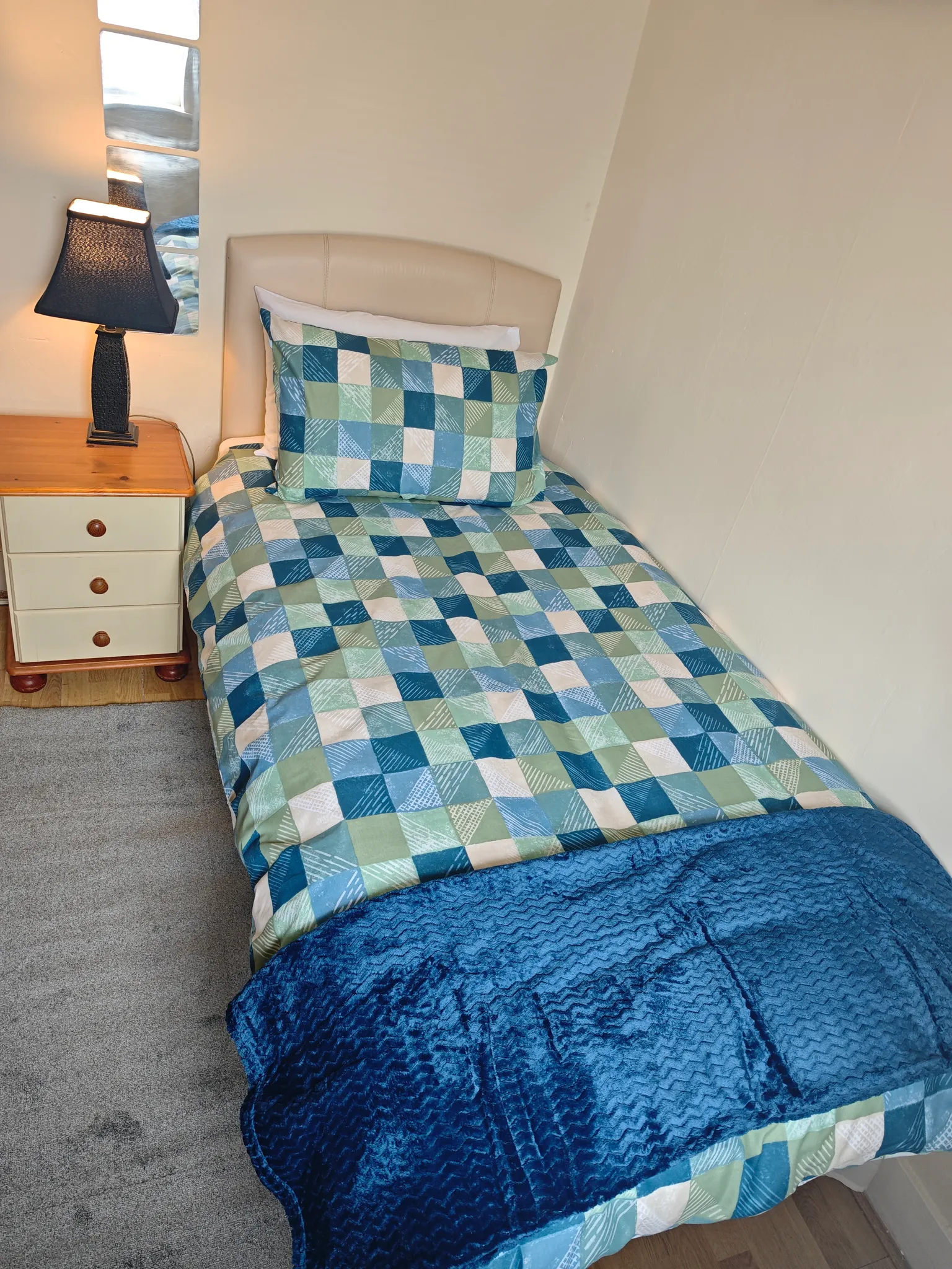 Bedroom with a single bed, patterned blue-green duvet and pillow, beige headboard, wooden bedside table with a lamp, and a small vertical mirror on the wall.