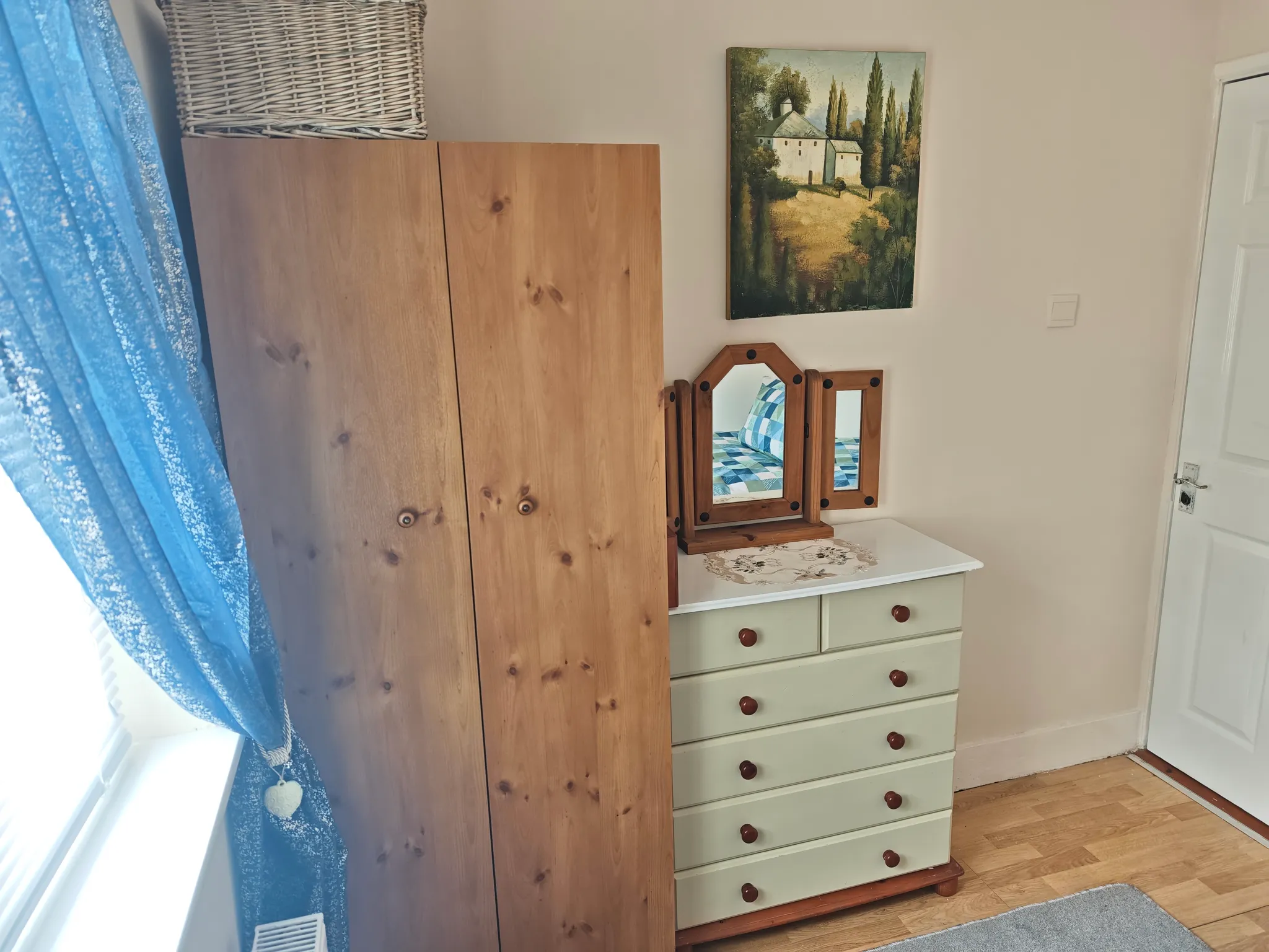 Bedroom corner with a wooden wardrobe, pale green chest of drawers topped by a tri-fold mirror, a blue curtain at a window, and a wall painting.