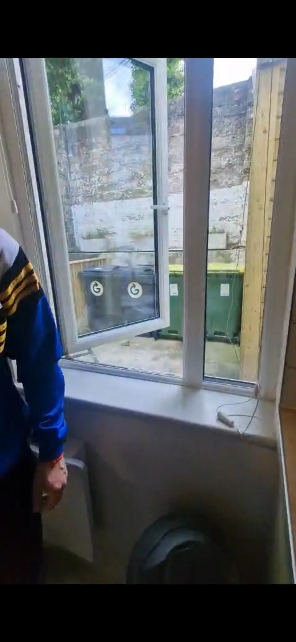 Room with a white open casement window and windowsill looking onto a small outdoor area with multiple trash bins and a stone wall.