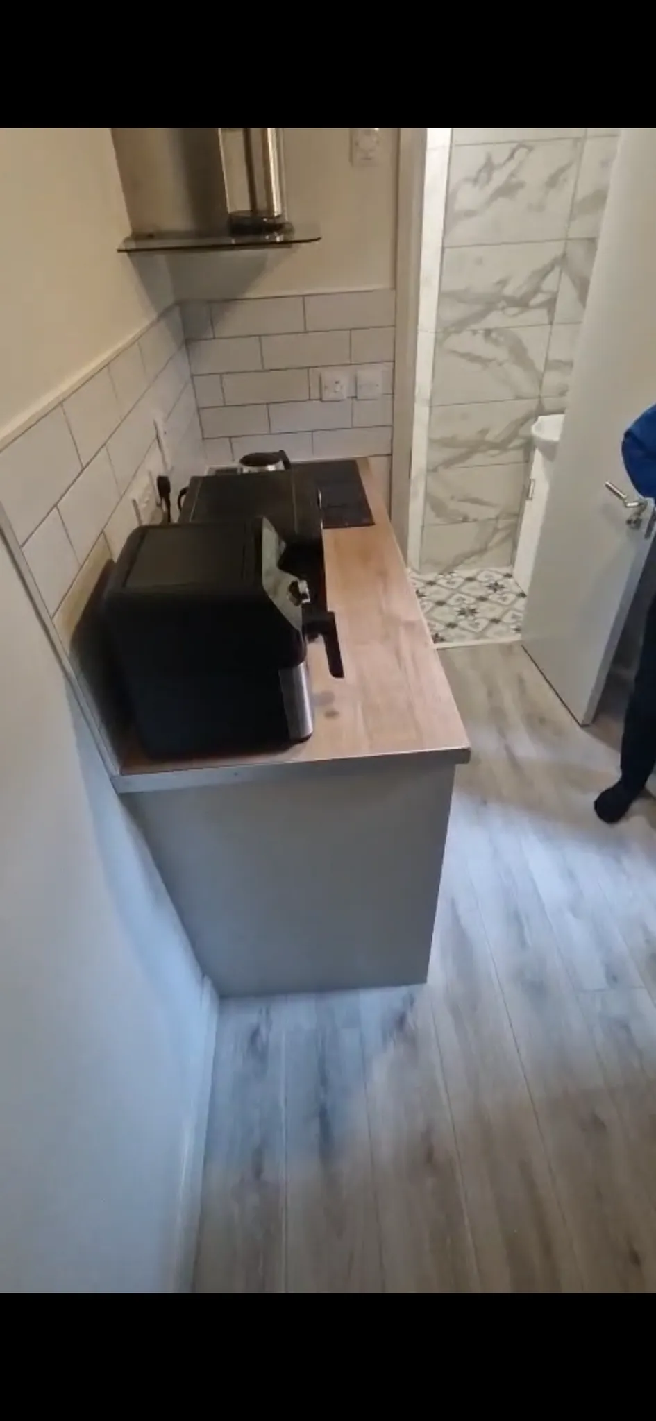 Small kitchen countertop with a toaster/air-fryer, electric hob, tiled backsplash and a stainless steel extractor hood; doorway to a tiled bathroom visible at the far end.