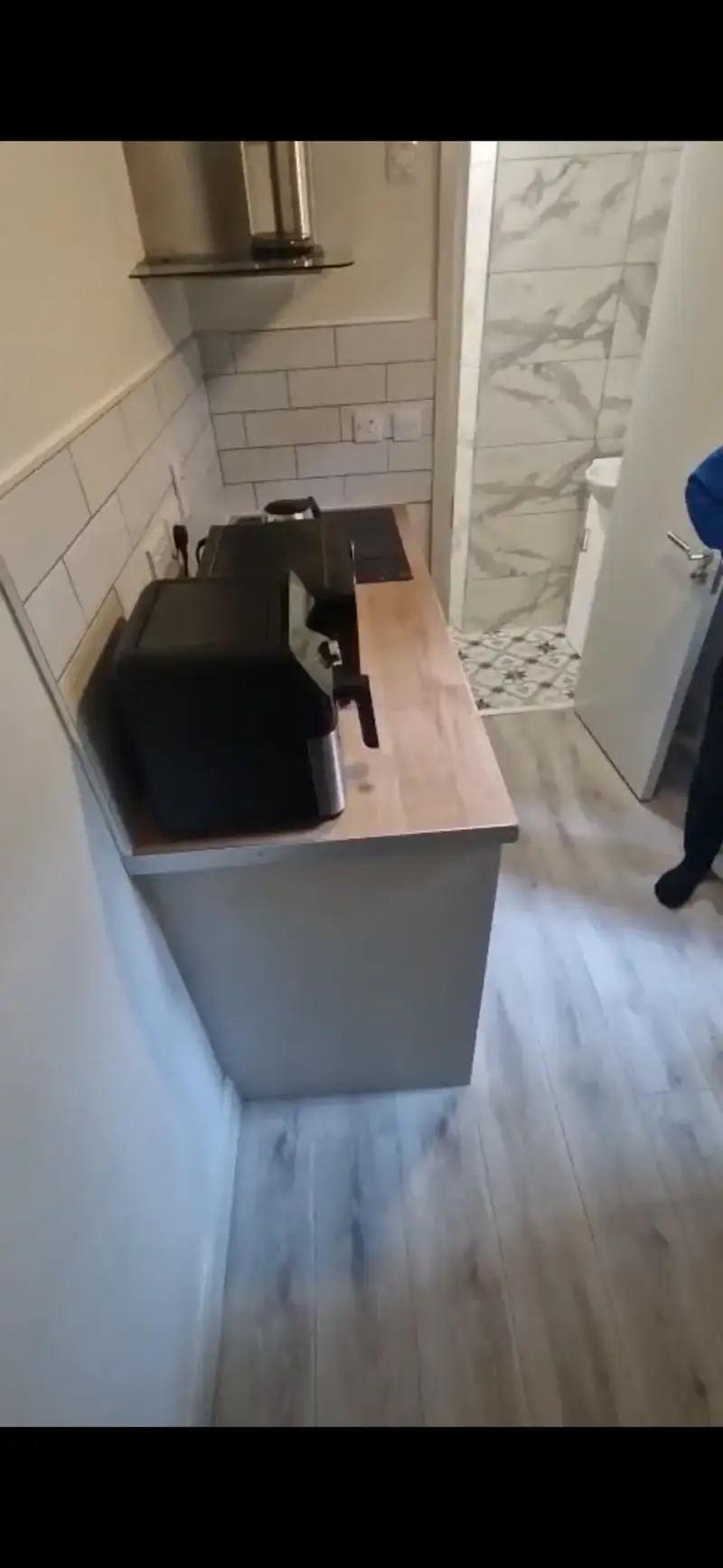 Small kitchen countertop with a toaster/air-fryer, electric hob, tiled backsplash and a stainless steel extractor hood; doorway to a tiled bathroom visible at the far end.