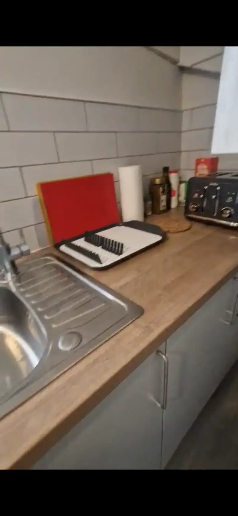 Kitchen countertop with a stainless steel sink and drainer, dish rack and cutting boards, paper towel roll, condiments and a toaster against a tiled backsplash.