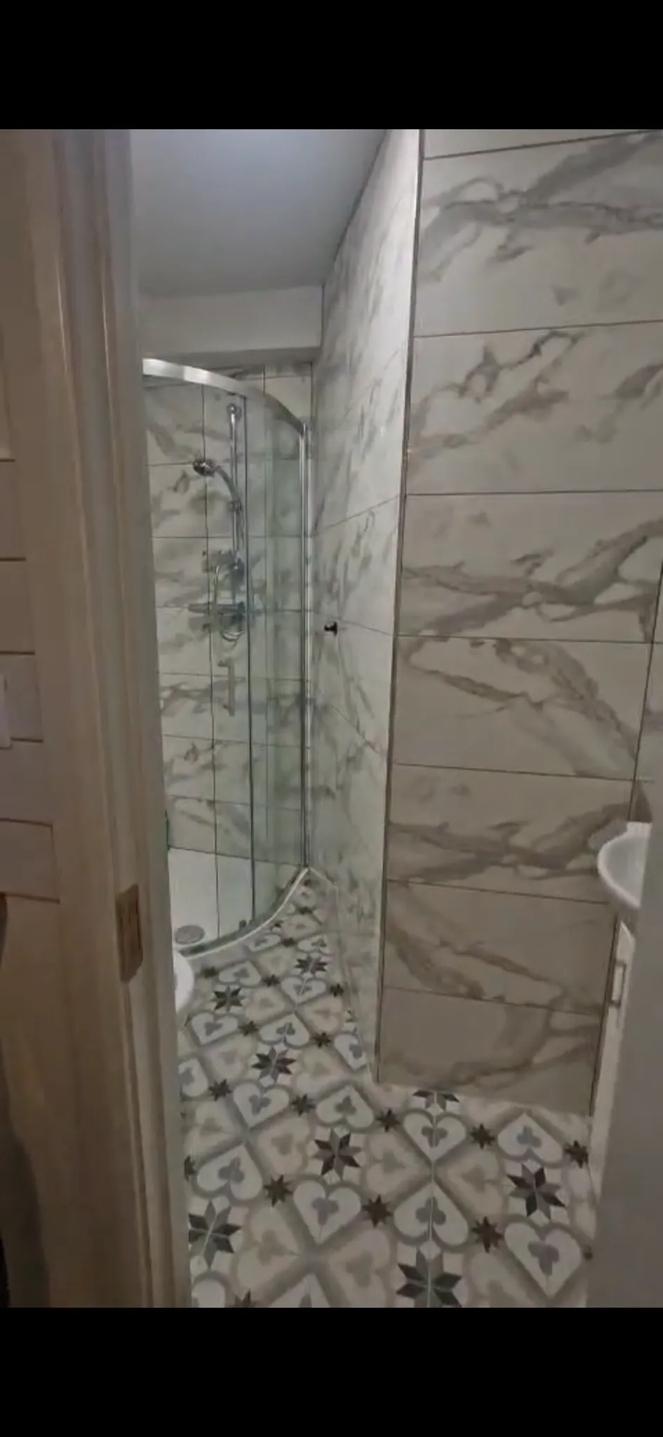 Bathroom with a curved glass corner shower, marble-look tiled walls and patterned floor tiles; a small sink is partially visible at right.