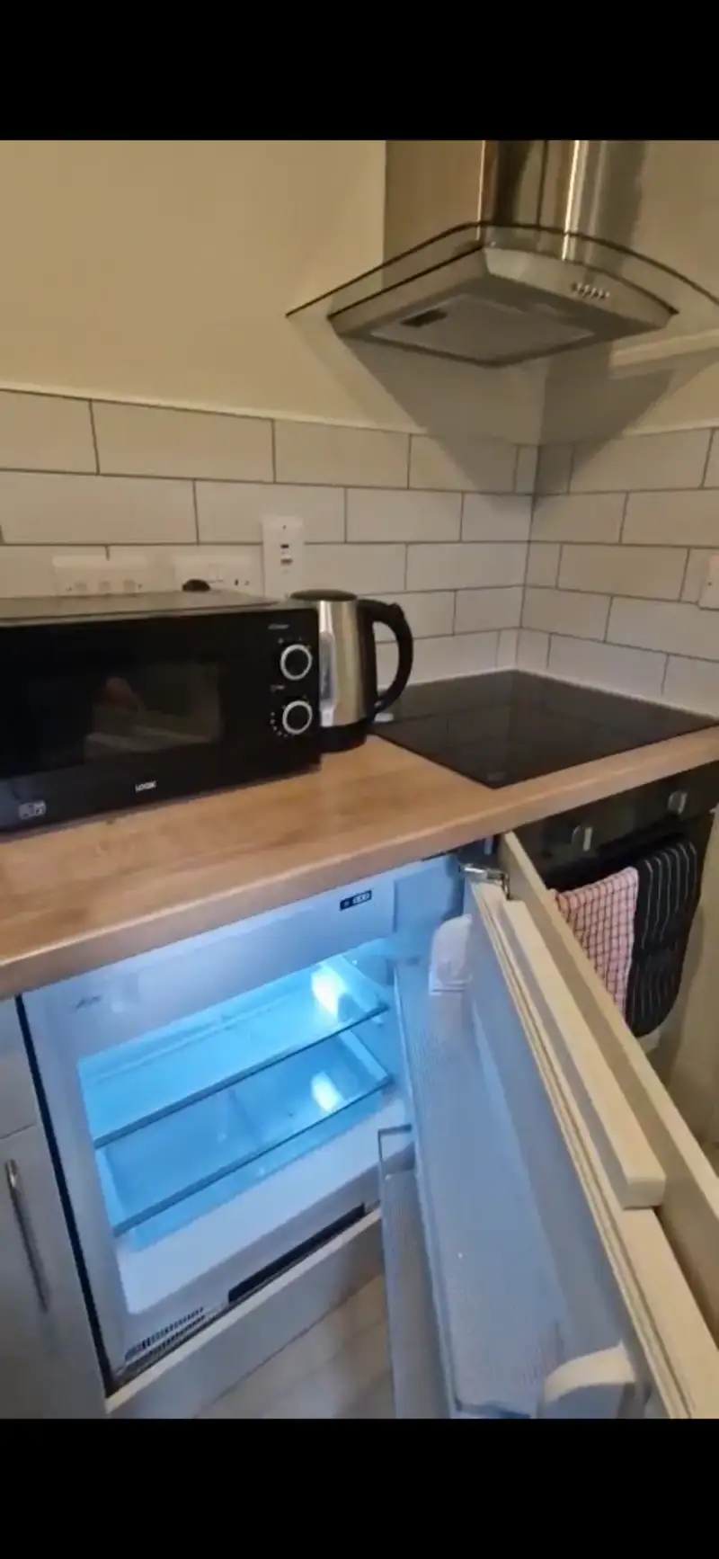Kitchen corner with a microwave, electric kettle, induction hob, extractor hood, and an open small fridge under a wooden countertop with tiled backsplash.