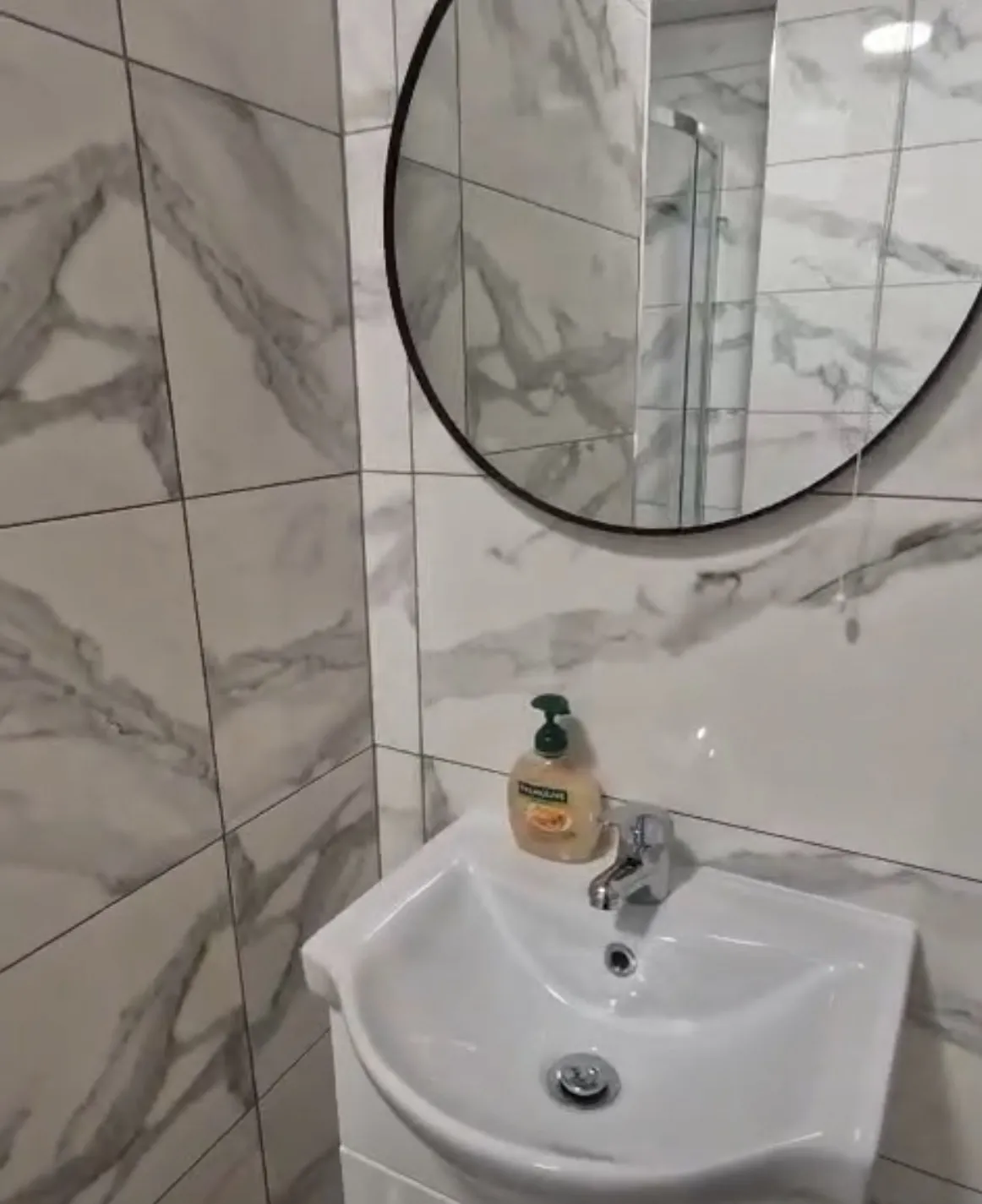 Bathroom with a small white sink, chrome faucet, round wall mirror, and soap dispenser on the sink; tiled marble-pattern walls.