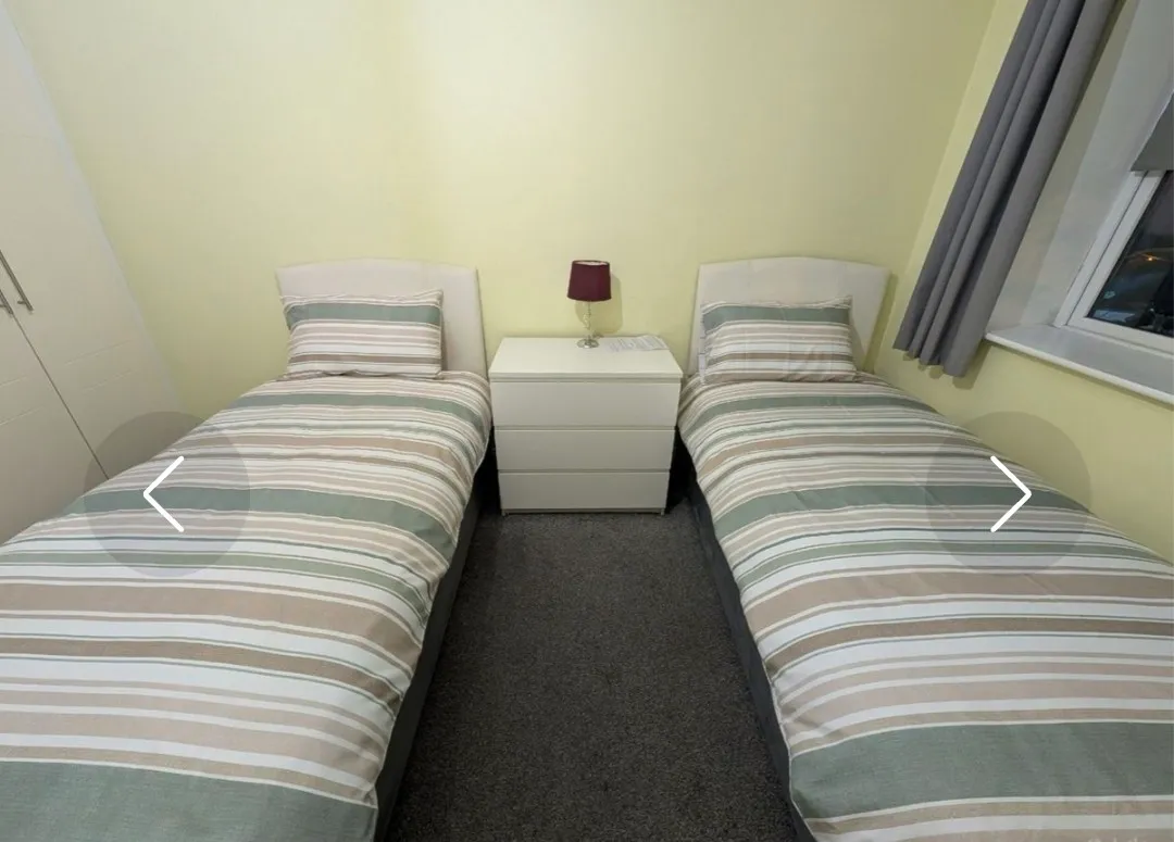 Bedroom with two single beds separated by a three-drawer nightstand holding a small table lamp; window with curtain and a wardrobe visible at left.