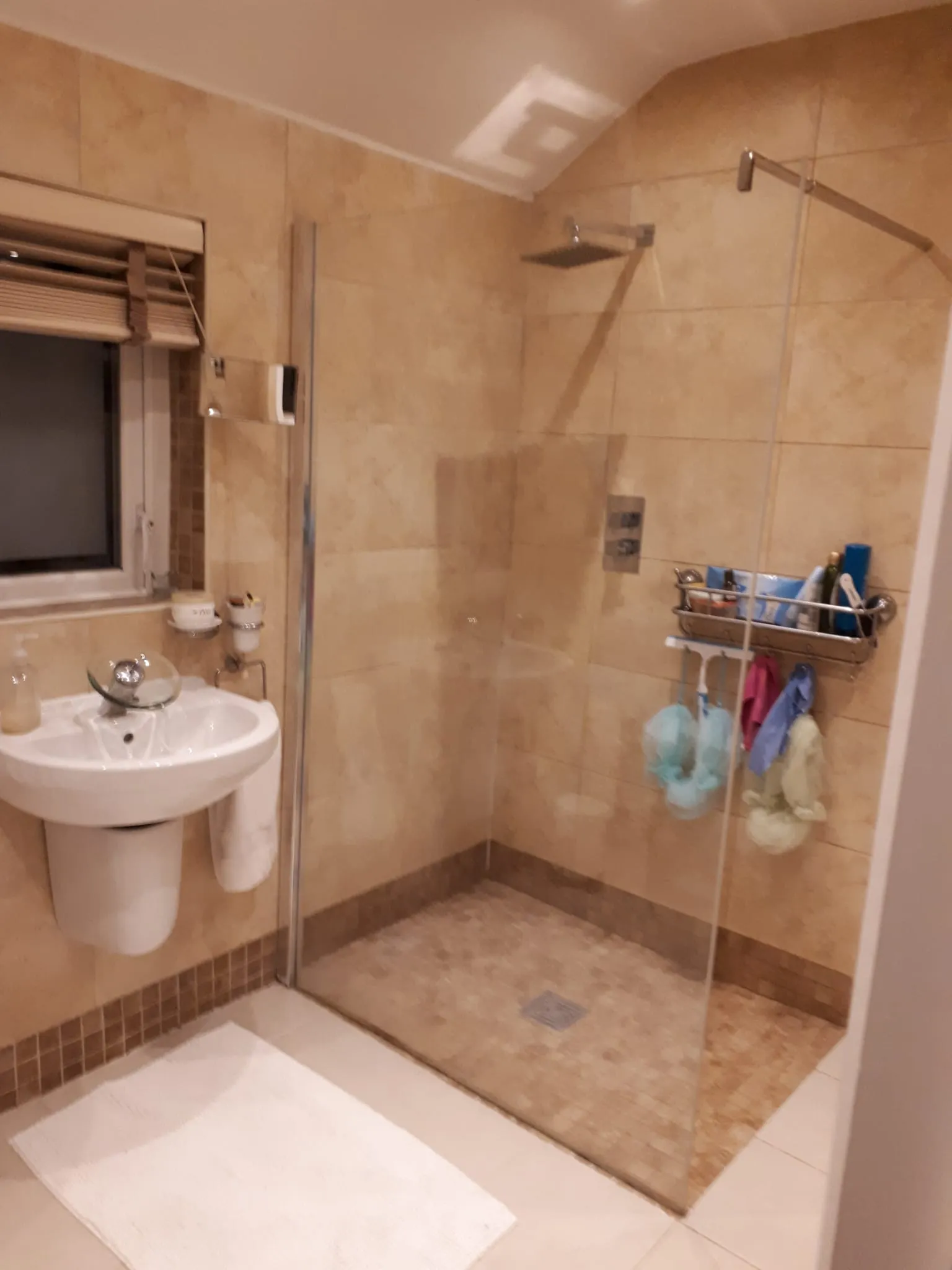 Bathroom with a walk-in tiled shower with glass partition, wall-mounted rain showerhead, pedestal sink and a small window.