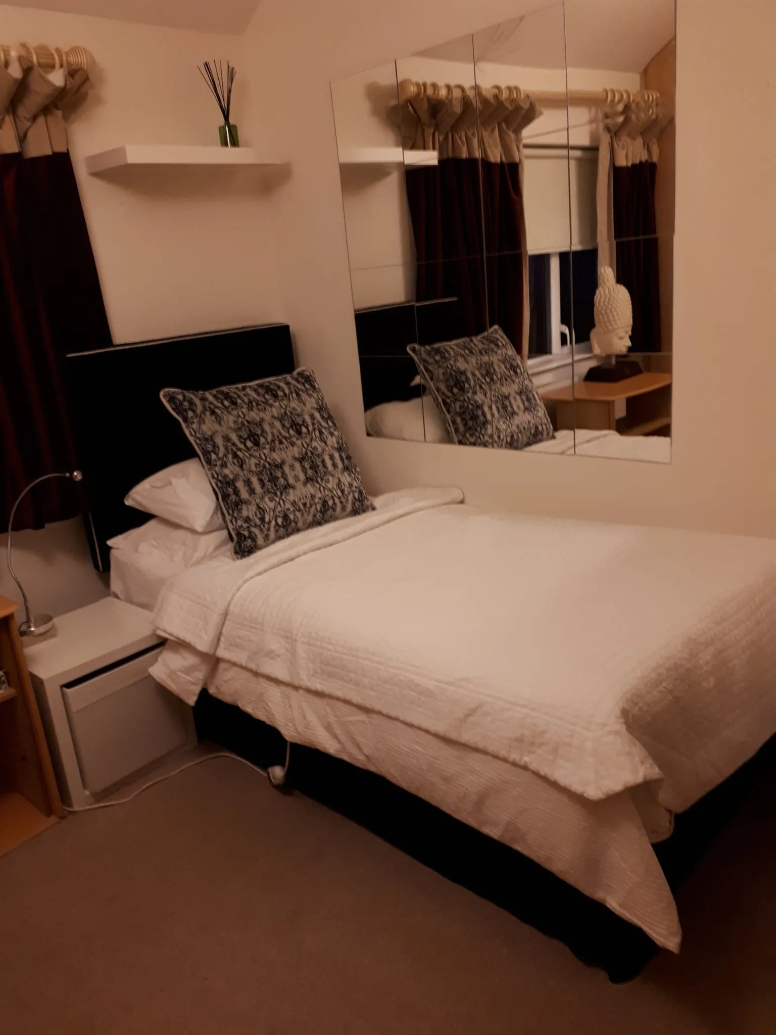Bedroom with a made bed, two patterned pillows, a mirrored wall above the bedside, curtains and a small bedside table with a decorative bust.
