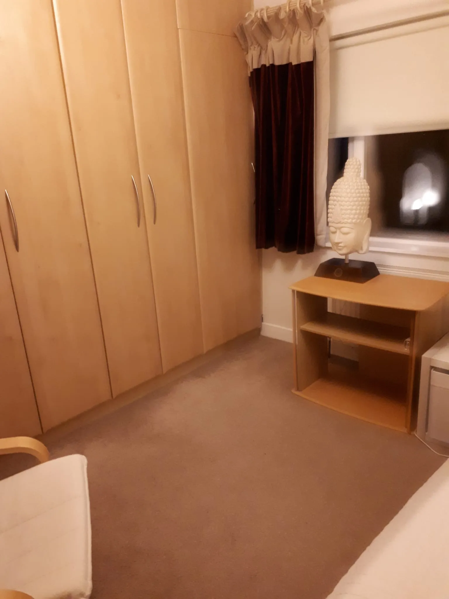 Bedroom corner with built-in wooden wardrobes, a window with dark curtains and a small shelving unit topped by a decorative head statue.