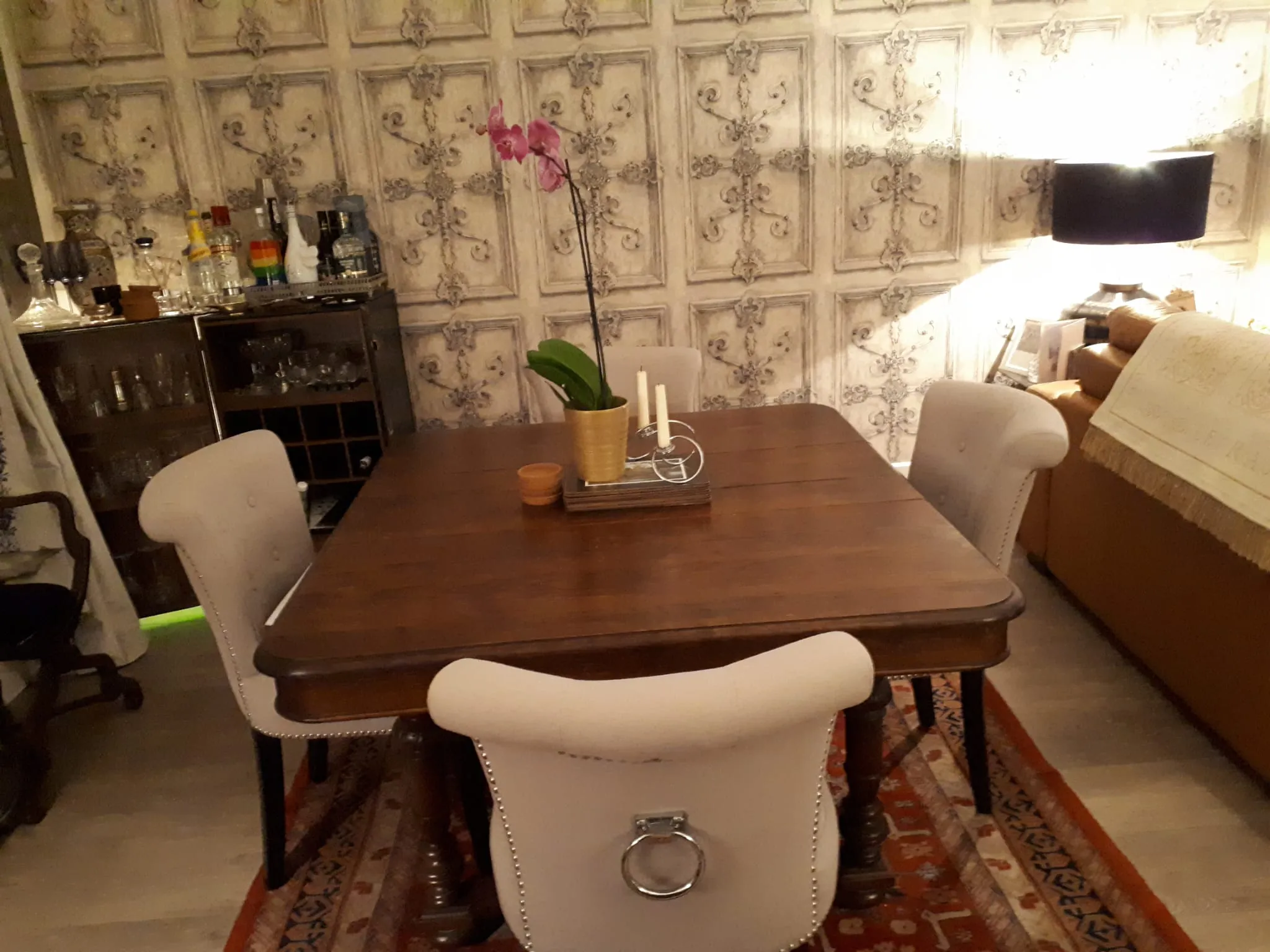 Dining room with a wooden dining table, upholstered chairs, a potted orchid and candlesticks on the table, patterned wall panels, a sideboard with glassware and a floor lamp.