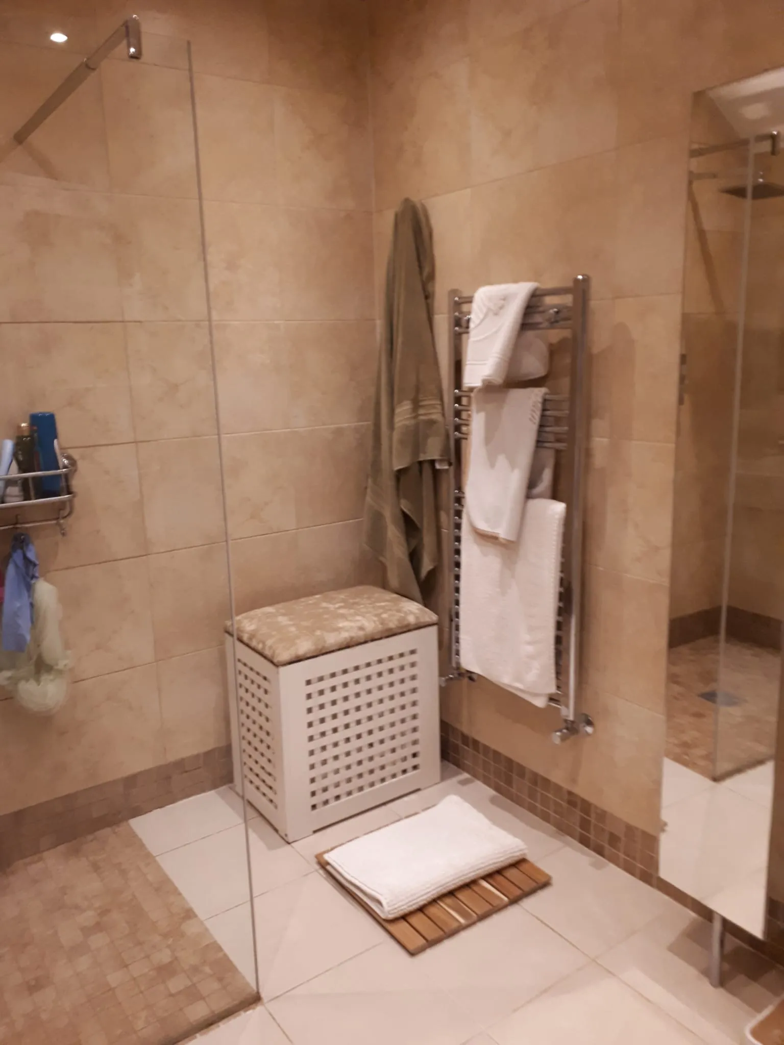 Bathroom with a walk-in shower, glass partition, wall-mounted heated towel rack with folded towels, laundry hamper and bath mat on tiled floor.