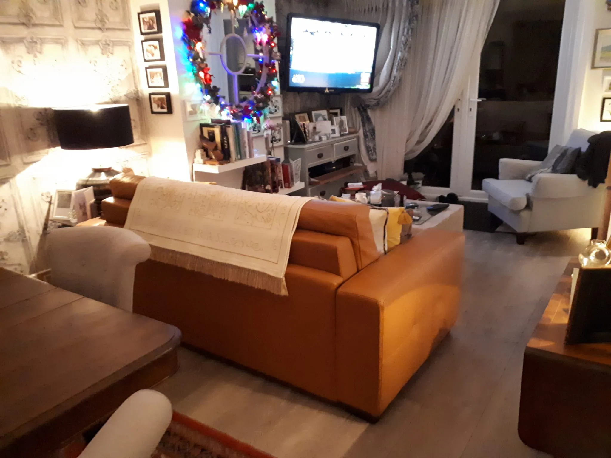Living room with a tan leather sofa, armchair, wall-mounted TV, coffee table and bookshelf with lit decorative wreath.