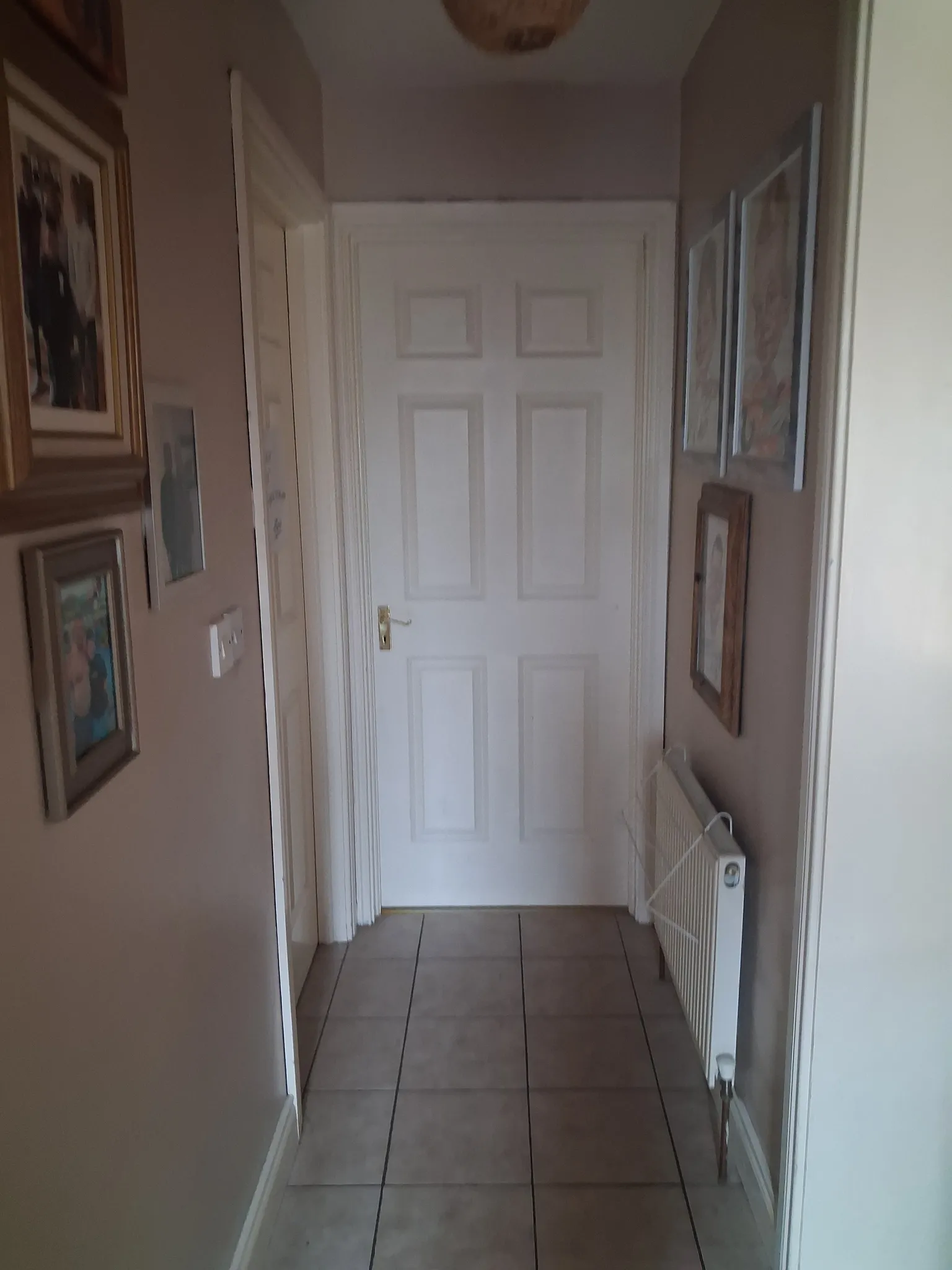 Narrow interior hallway with a closed white paneled door at the end, tiled floor, wall-mounted radiator on the right and framed pictures on both walls.