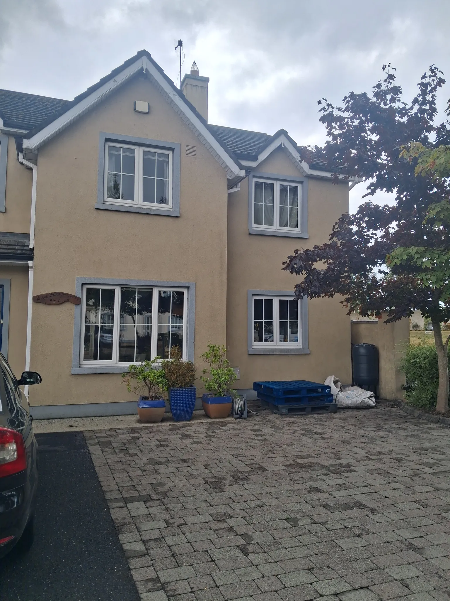 Exterior view of a two-story house facade with multiple windows, a paved driveway, potted plants near the entrance and a tree at right.