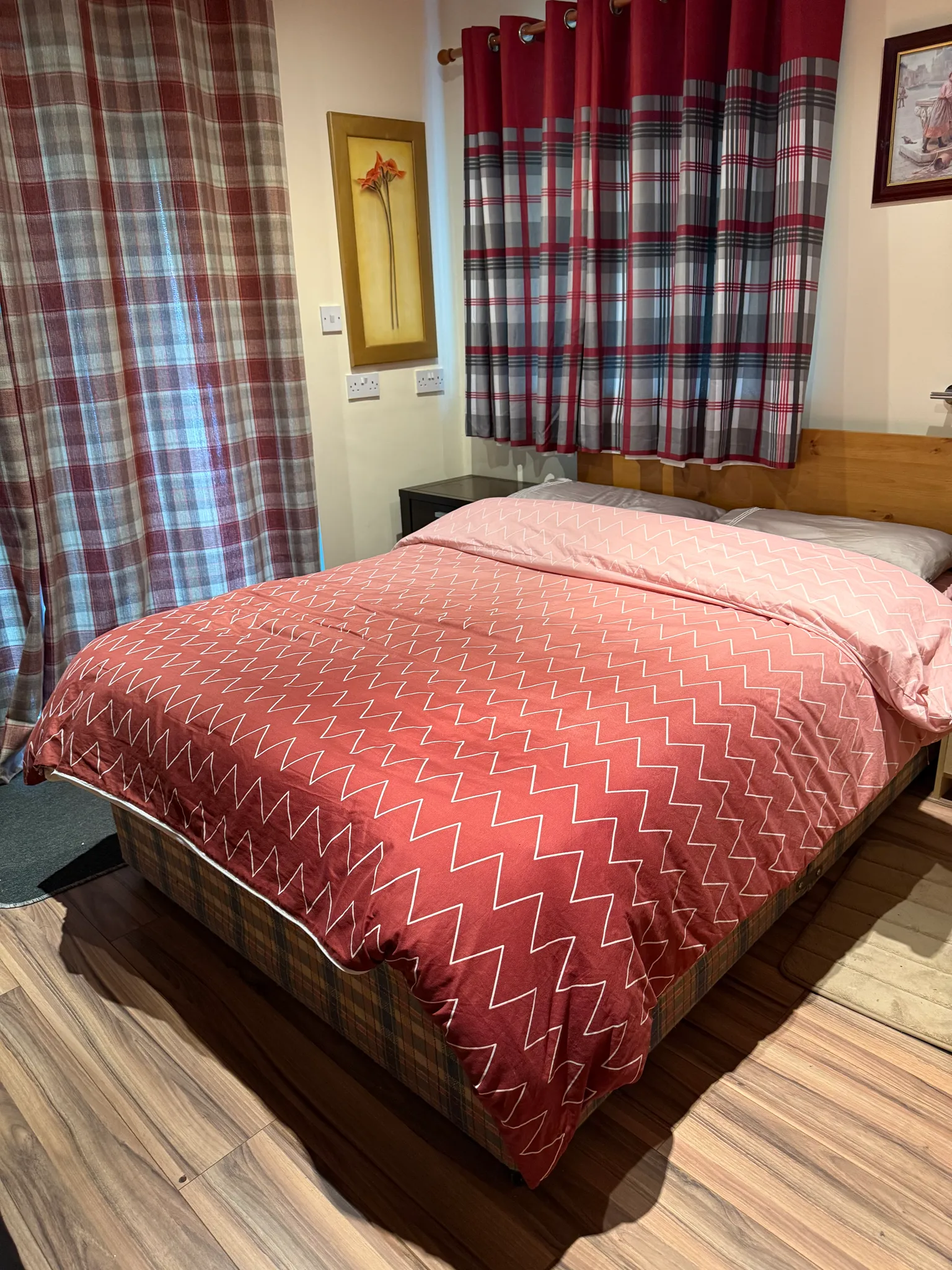 Bedroom with a double bed covered by a patterned red duvet, plaid curtains, a small bedside table and wall art.