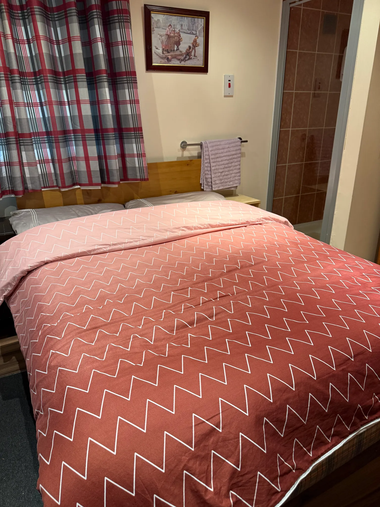 Bedroom with a double bed and pink patterned duvet, plaid curtains, framed picture on the wall, towel on a wall rail and a doorway to a tiled bathroom.