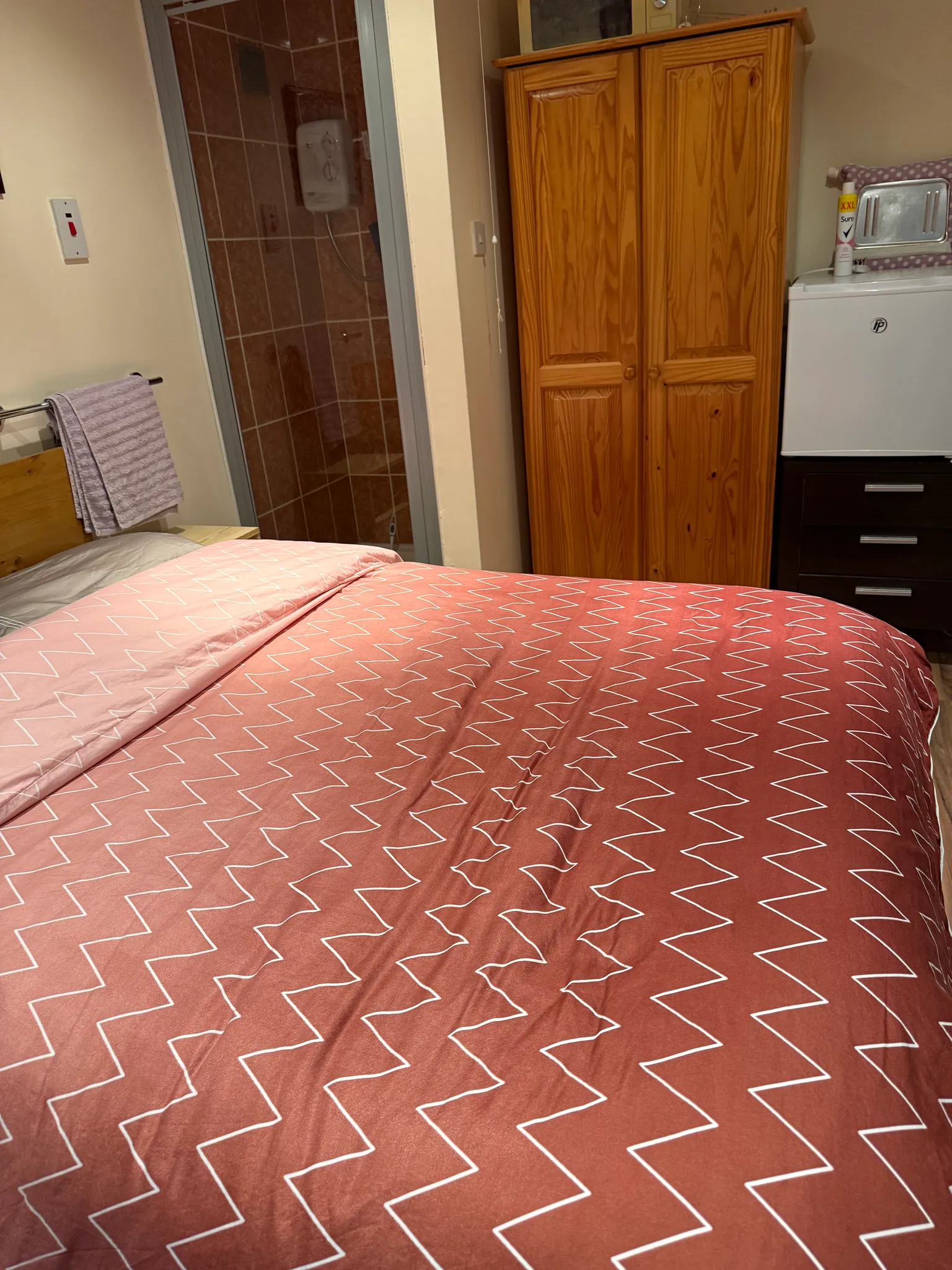 Bedroom with a double bed in a patterned duvet, wooden wardrobe, small fridge on a chest of drawers and a tiled shower cubicle visible through a doorway.