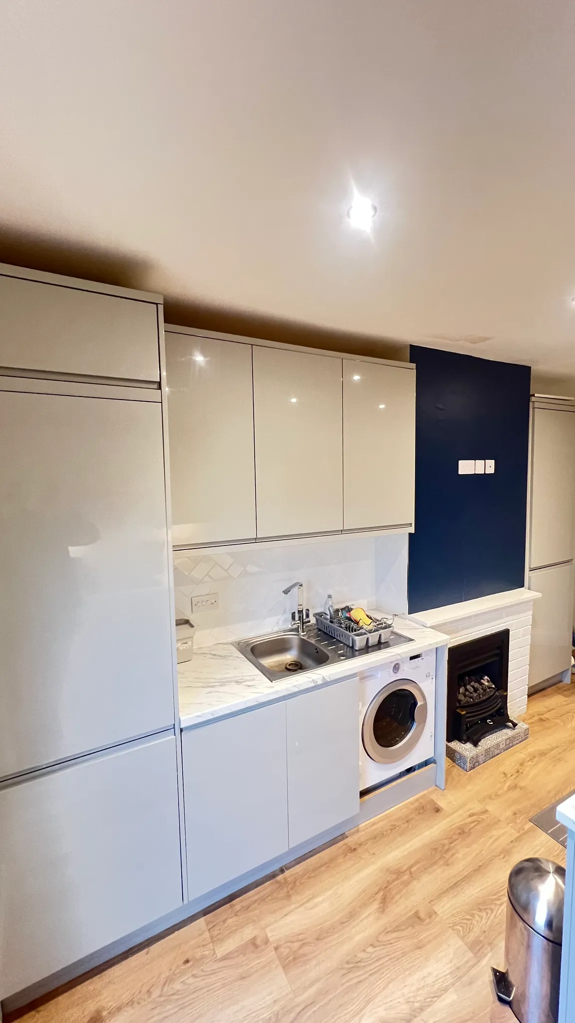 Kitchen with upper and lower cabinets, a sink with dish rack, a front-loading washing machine under the counter, a small fireplace, and wood-effect floor.