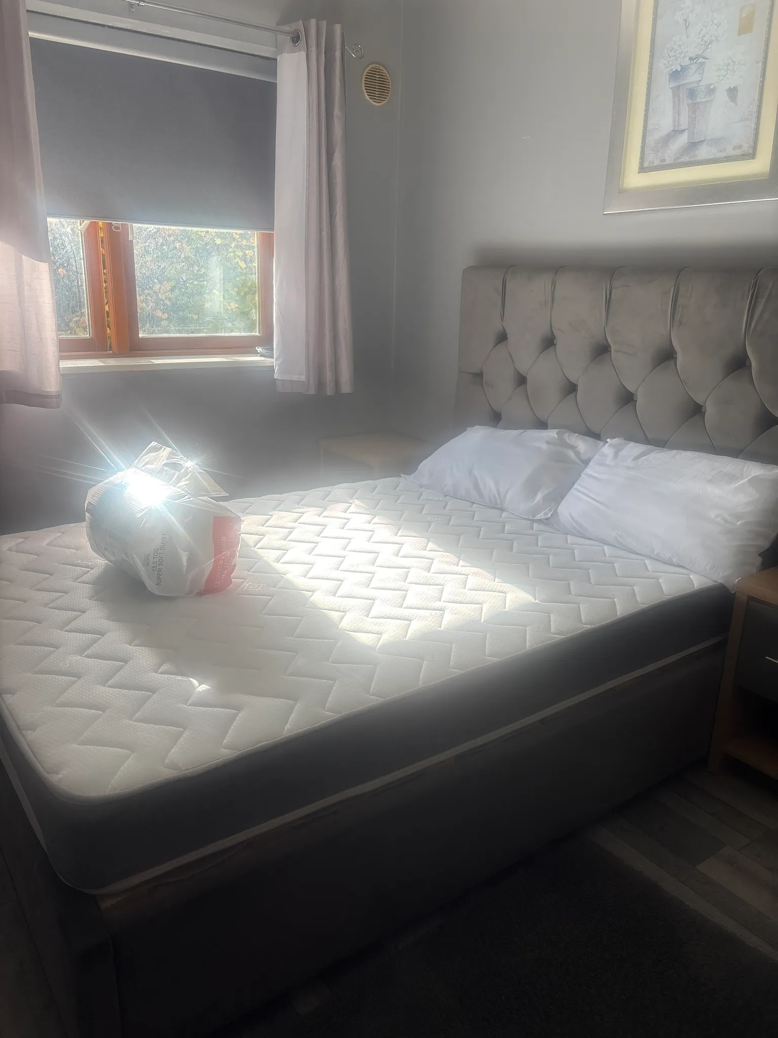 Bedroom with a mattress on a bed frame, tufted headboard, two pillows, a window with curtain and roller blind, and sunlight on the mattress.