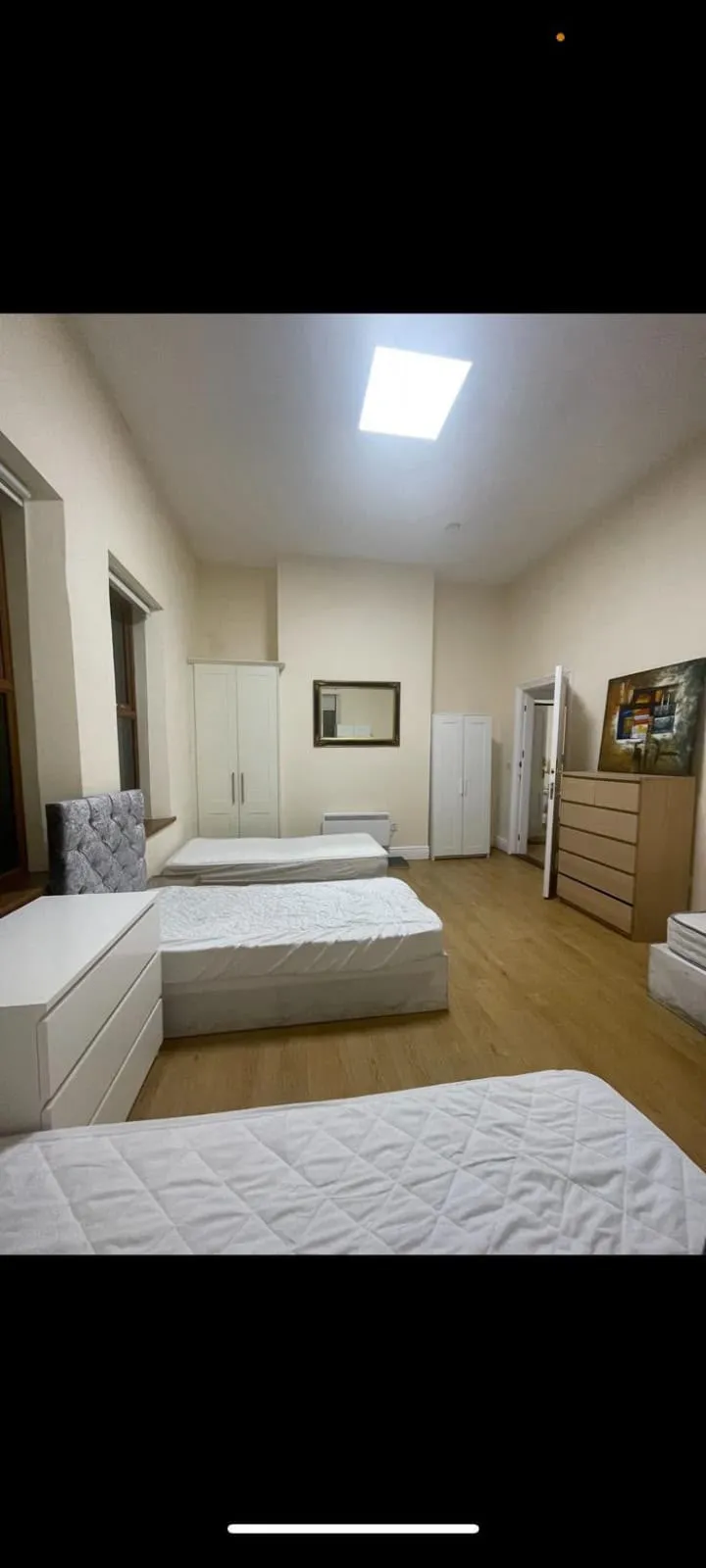 Bedroom with three single beds, two white wardrobes, a tall chest of drawers, a wall mirror, and a ceiling light panel.