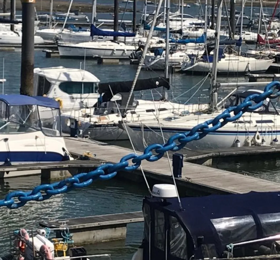 Marina with multiple moored sailboats and docks, large blue chain crossing the foreground over the water.
