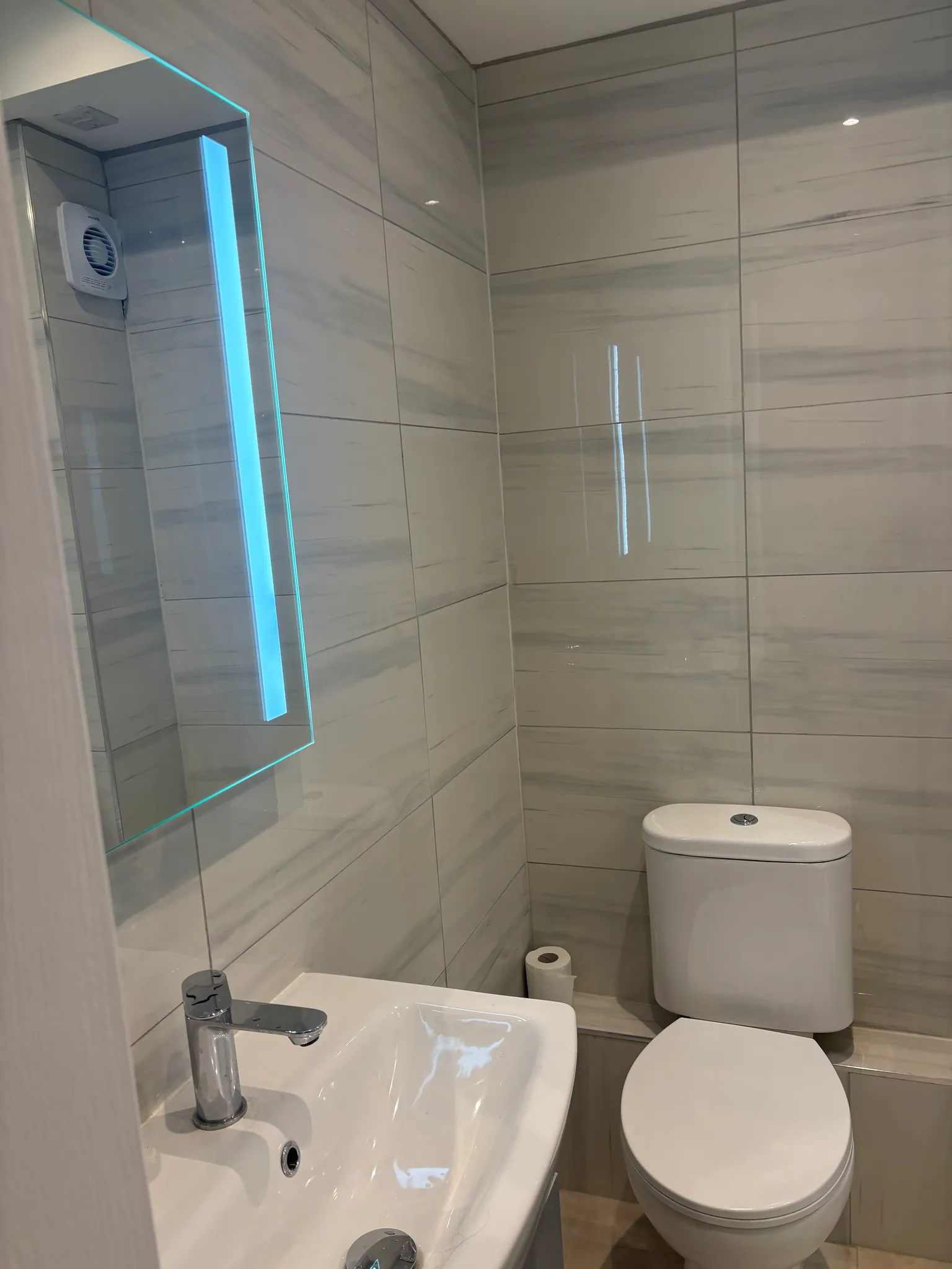 Small bathroom with a toilet, sink with chrome faucet, illuminated mirror, and tiled walls.