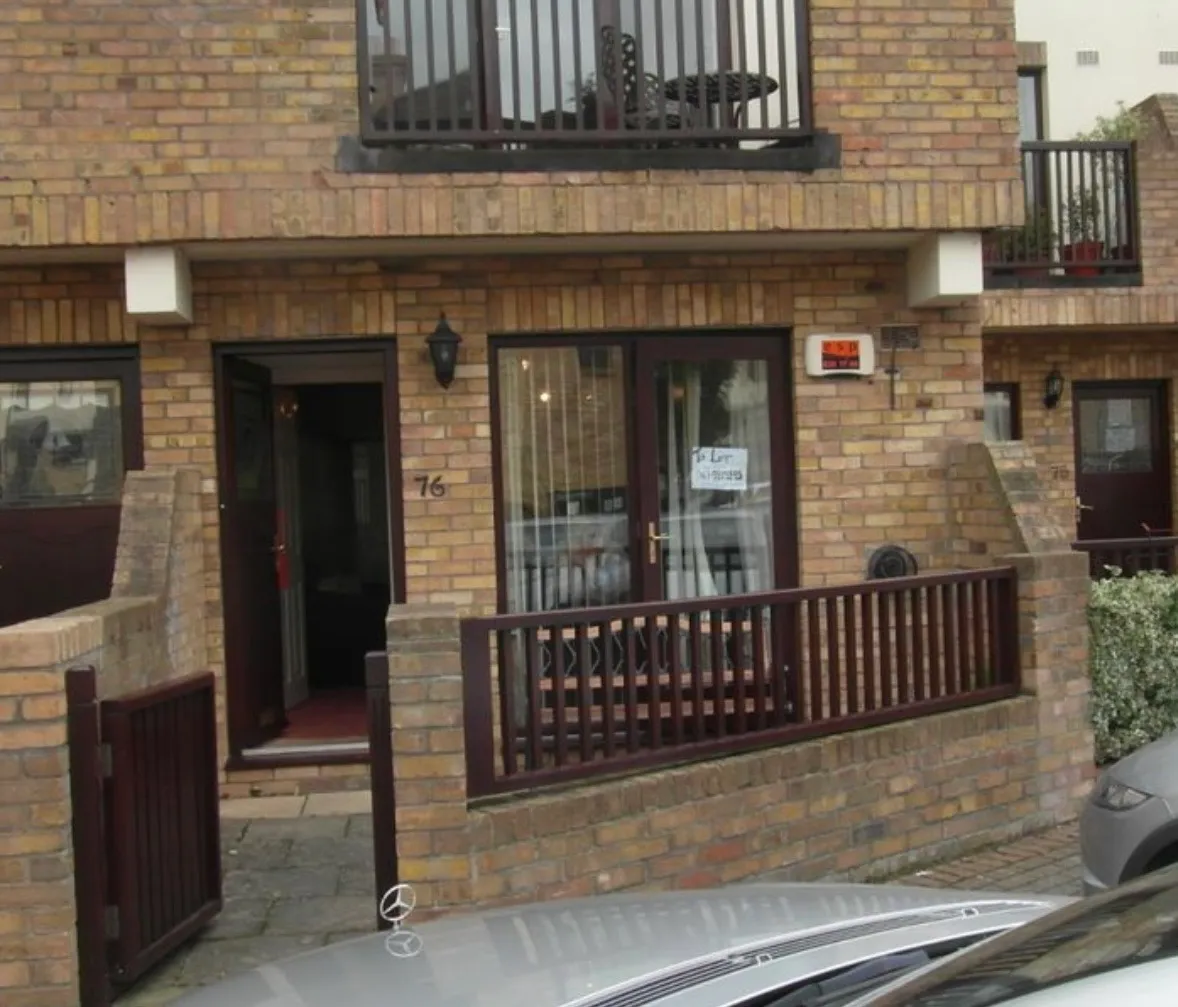 Ground-floor brick building facade with a small gated patio, front door and glass patio doors showing a 'To Let' sign, and a balcony above.