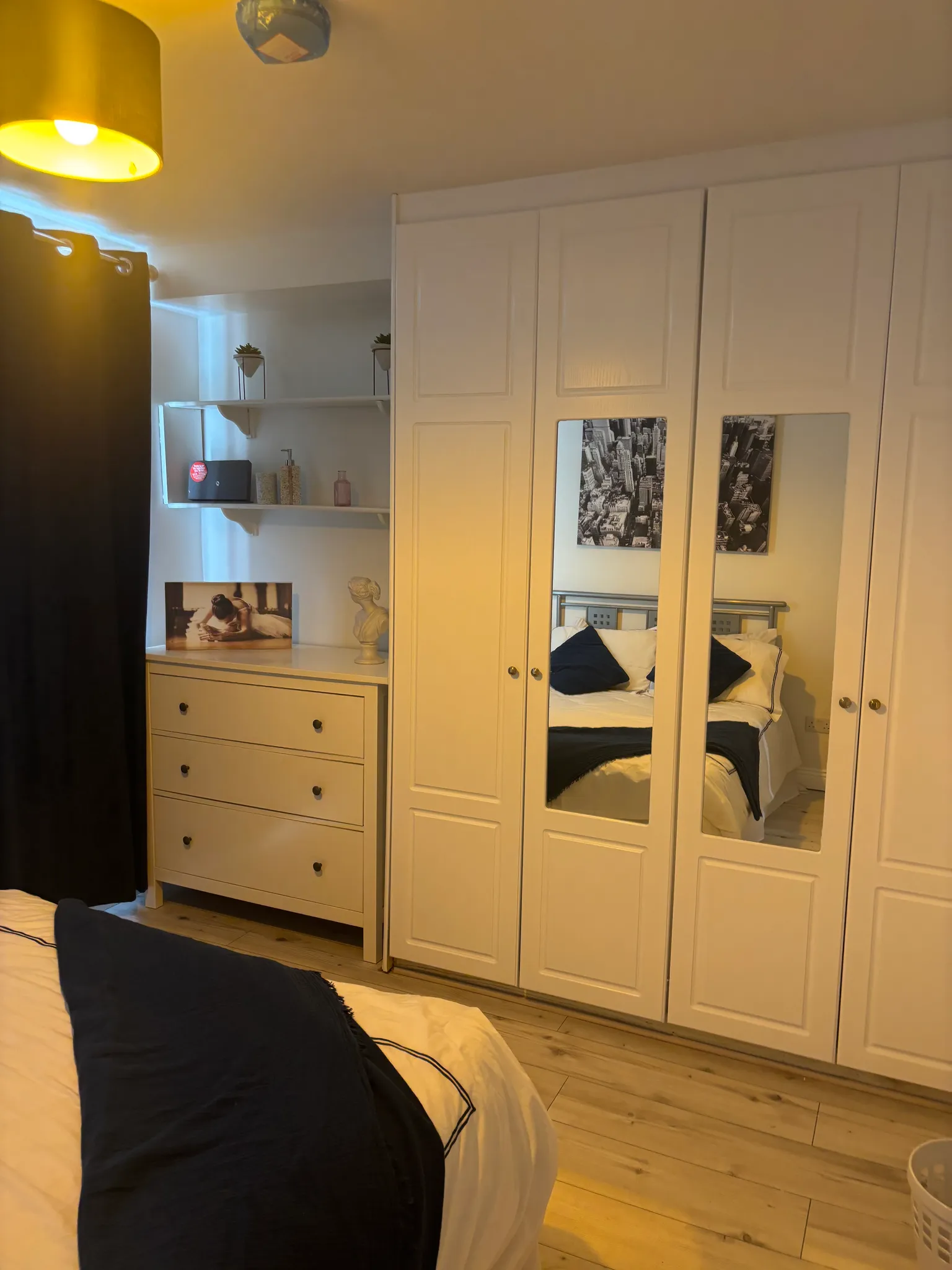 Bedroom showing a bed with pillows, a tall white wardrobe with mirrored doors, a chest of drawers and shelving with decor.