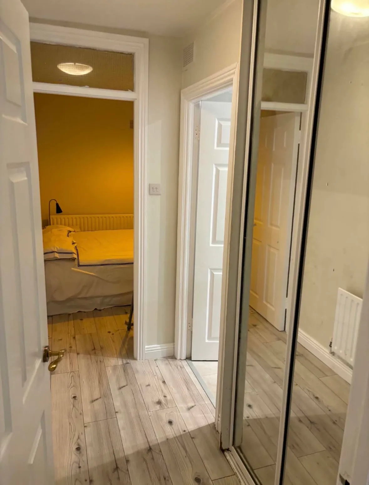 Hallway with wood-look floor, mirrored sliding wardrobe doors on the right, open doorways leading to a bedroom with a bed and bedside lamp.