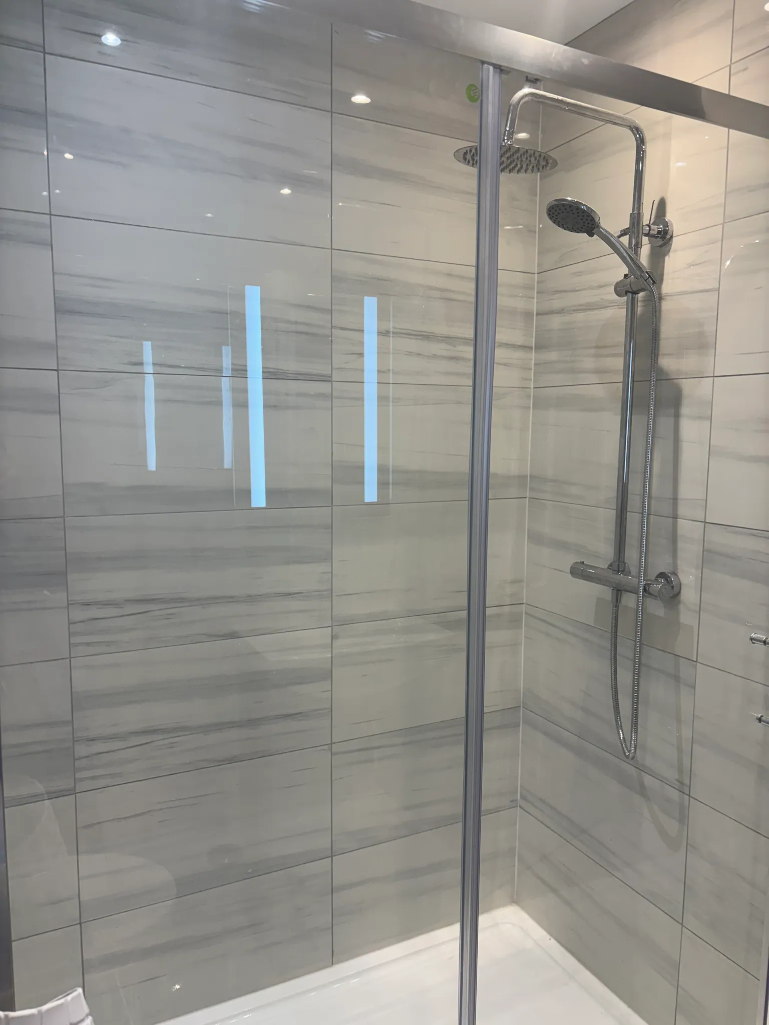 Bathroom shower stall with glass door, tiled walls, a wall-mounted rainfall showerhead and detachable handheld shower on chrome fittings.
