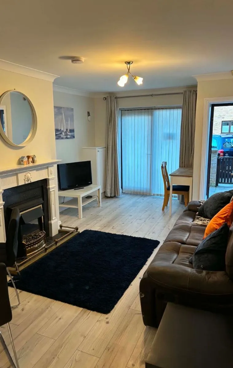 Living room with a brown leather sofa, black rug on light wood floor, fireplace with mantel and a small TV on a white stand, sliding glass door with vertical blinds and a small dining table by the door.