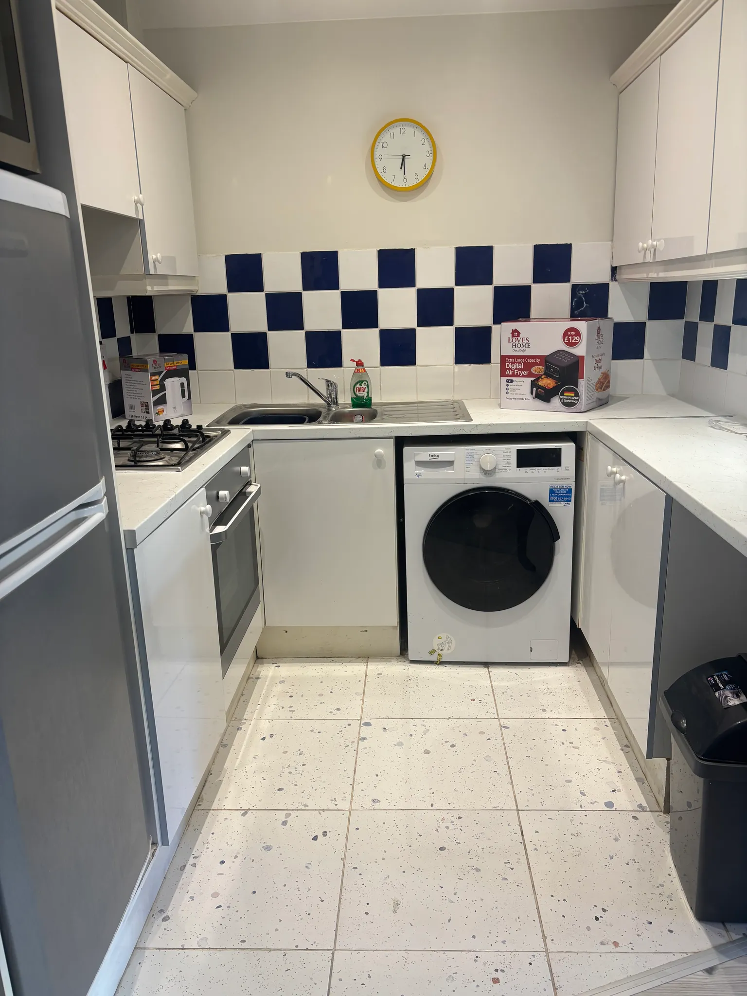 Small kitchen with white cabinets, a sink, gas hob and oven, washing machine under the counter, blue-and-white tiled backsplash and a yellow-rimmed wall clock