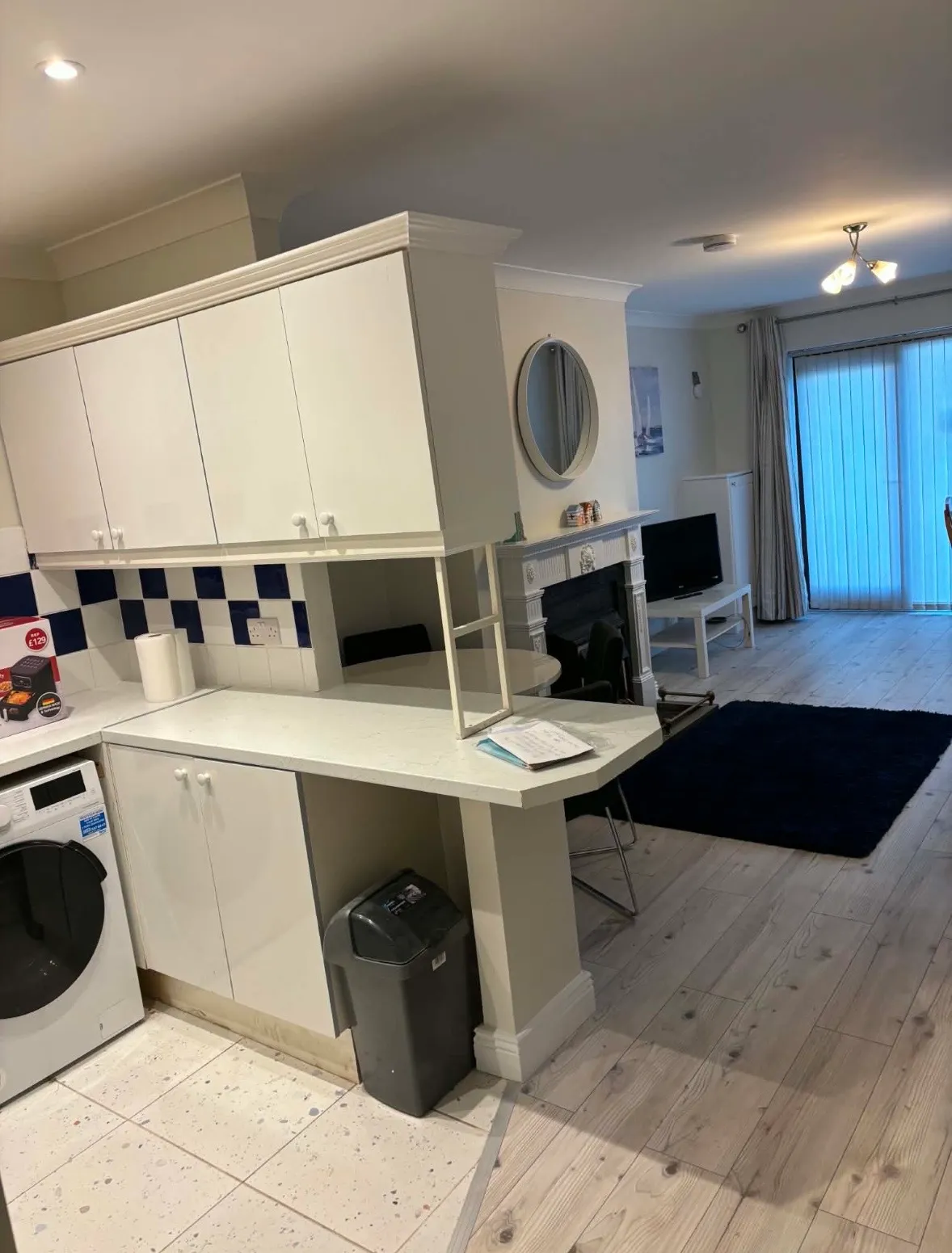 Open-plan living area showing a kitchen counter with white cabinets and a washing machine, a small dining counter, fireplace with round mirror above, TV on a stand, rug, and sliding glass door with vertical blinds.