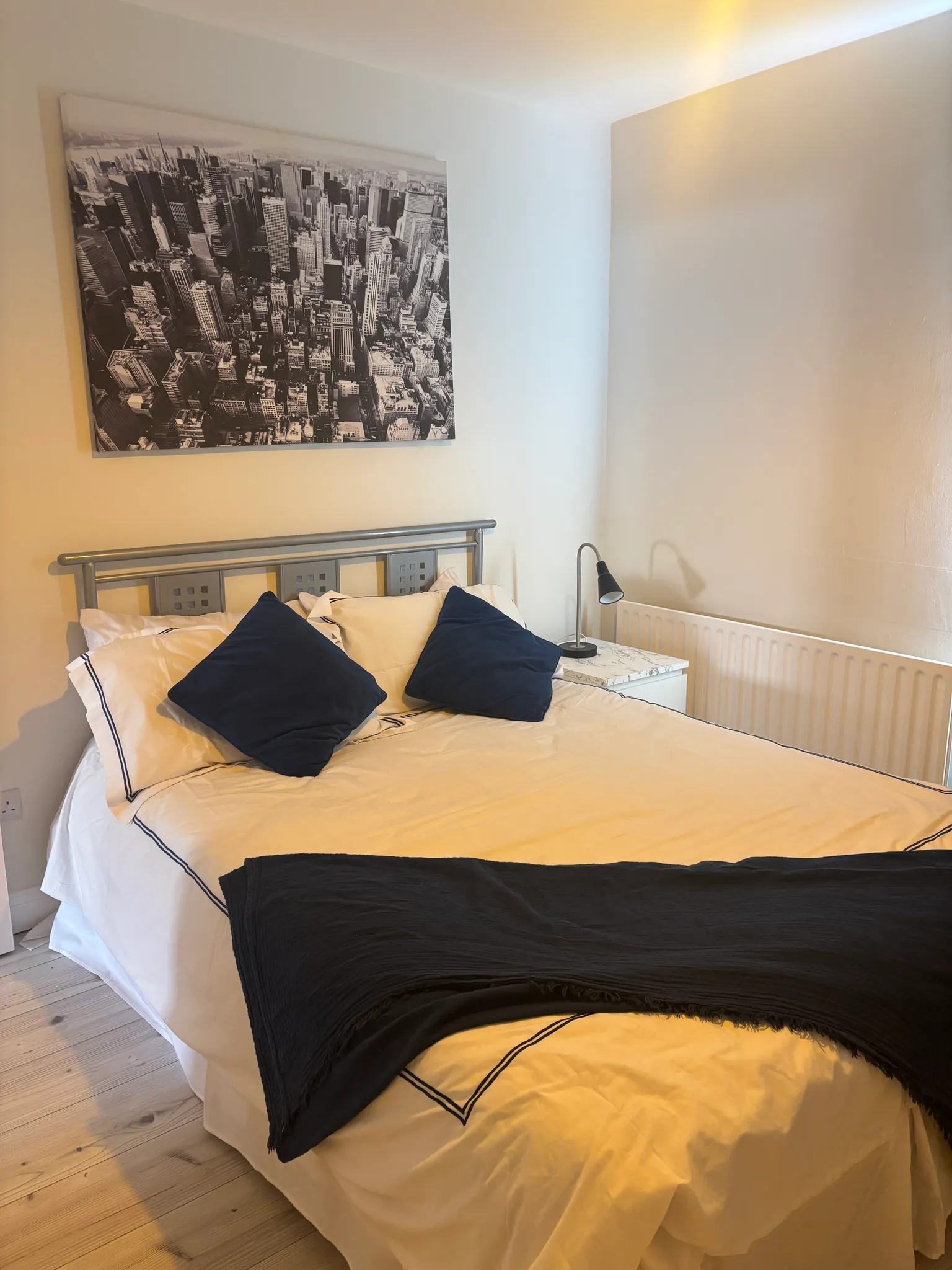 Bedroom with a double bed, two dark pillows, a headboard, bedside table with a lamp, radiator and black-and-white city print on the wall.