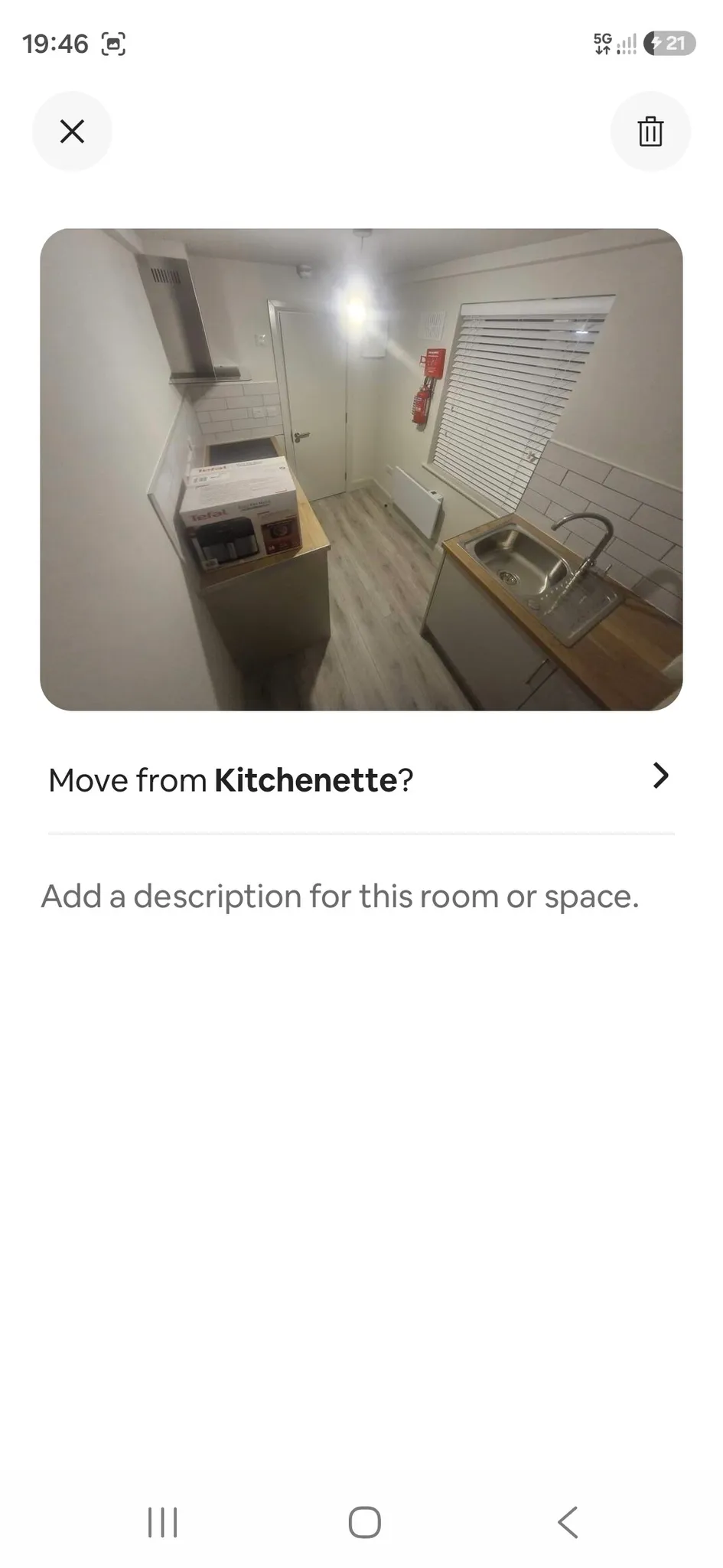 Small kitchenette-style kitchen with a sink and wooden countertop on the right, a counter with a boxed appliance on the left, window with blinds, radiator and a closed door at the far end.