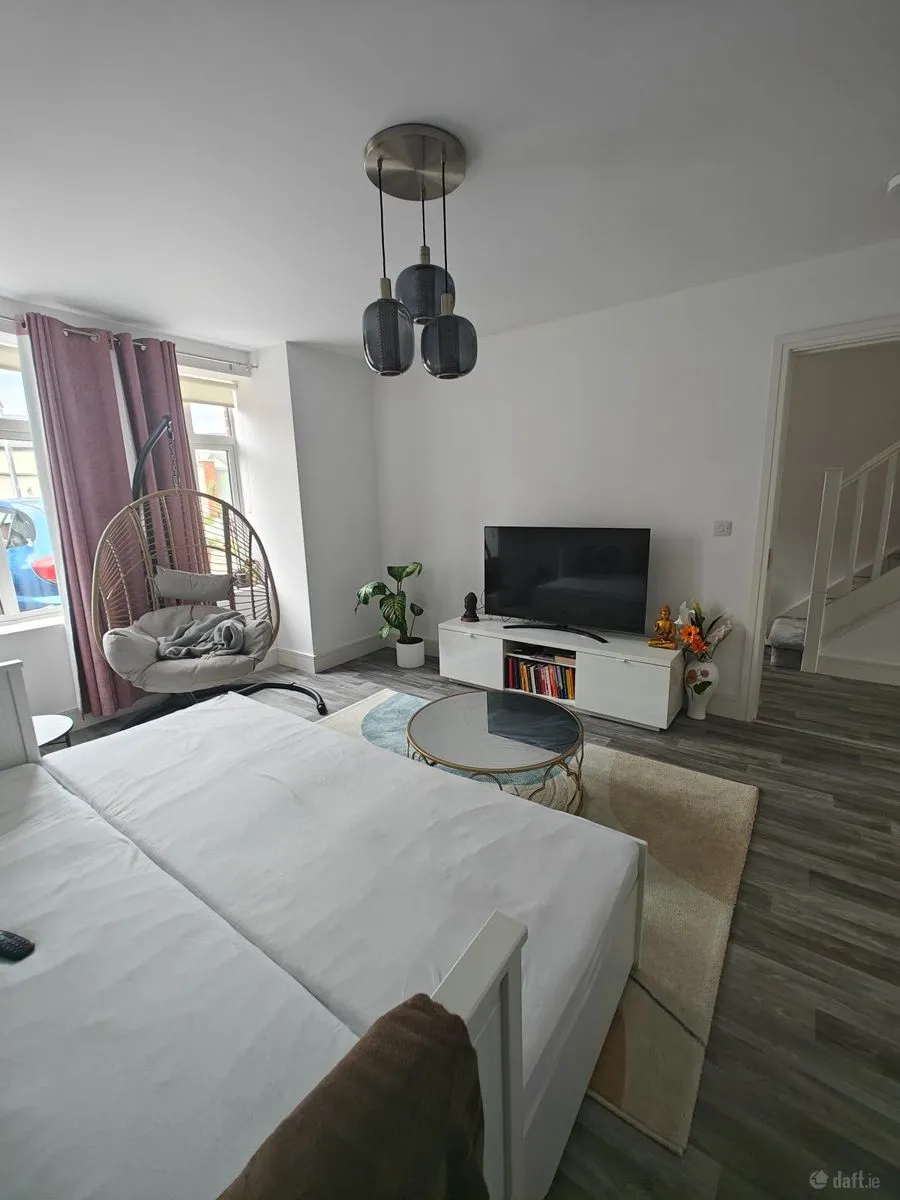 Bedroom with a double bed, wall TV on a low media console, round glass coffee table, hanging rattan chair by a window with curtains, potted plant, and pendant ceiling lights.