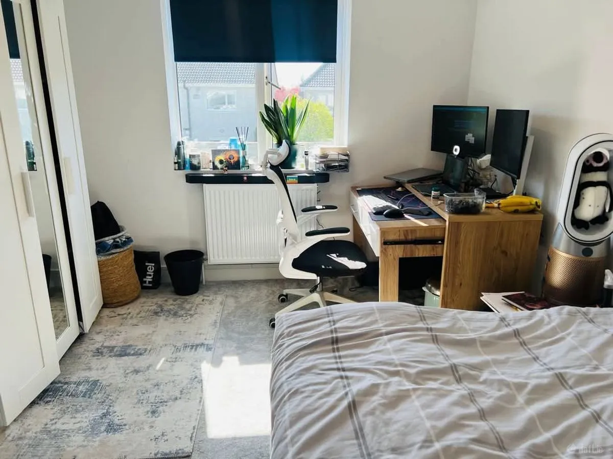 Bedroom with bed in foreground, desk with dual monitors and white office chair positioned by a window with a roller blind.