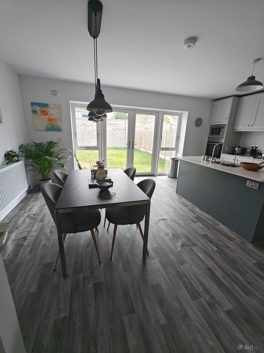 Open-plan dining area and kitchen with a rectangular dining table and four chairs, kitchen island with sink, pendant lights, and glass patio doors opening to a small lawn.