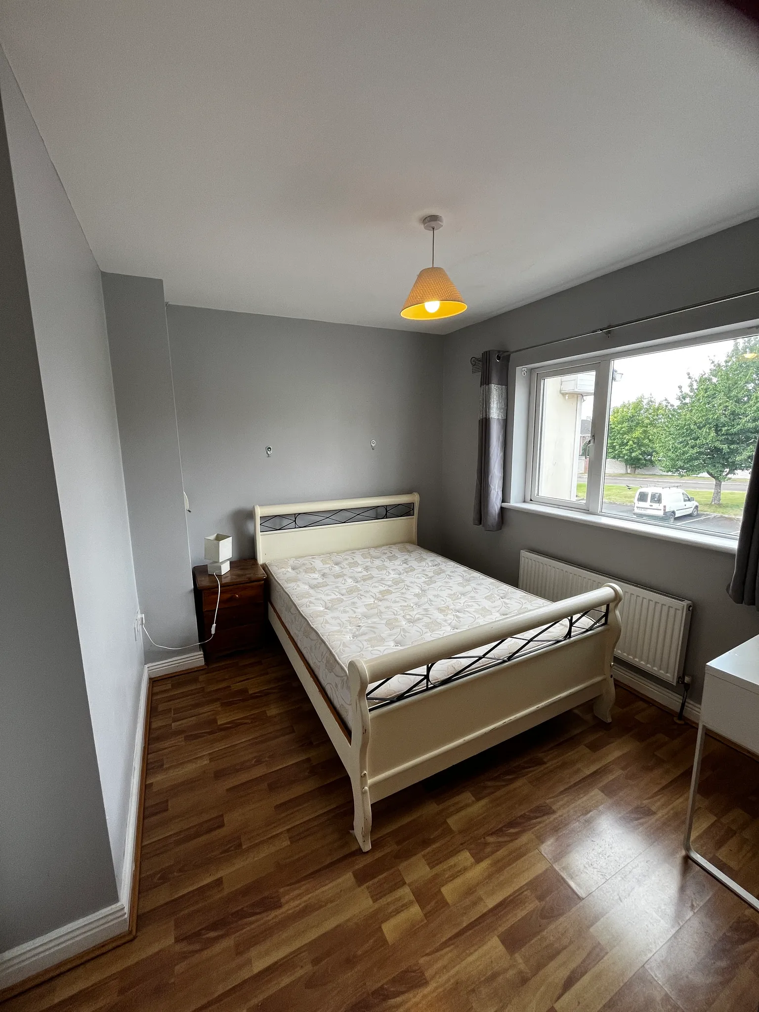 Bedroom with a double bed, bedside table and lamp, window with curtains, radiator and ceiling pendant light.