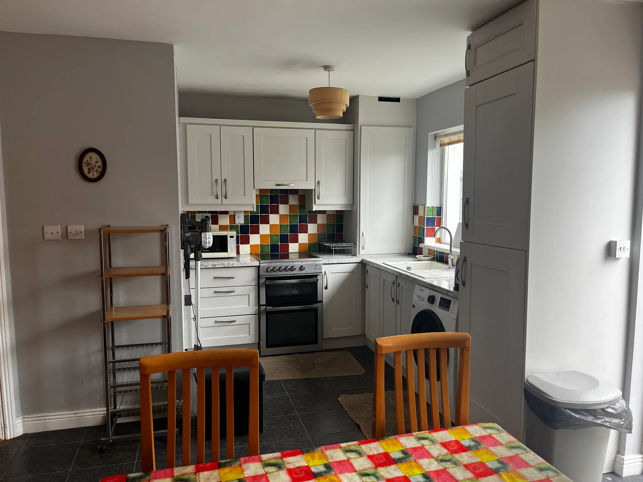 Kitchen with white cabinets and multicolored tile backsplash, freestanding oven, countertop sink under a window, pendant light and dining chairs in foreground.