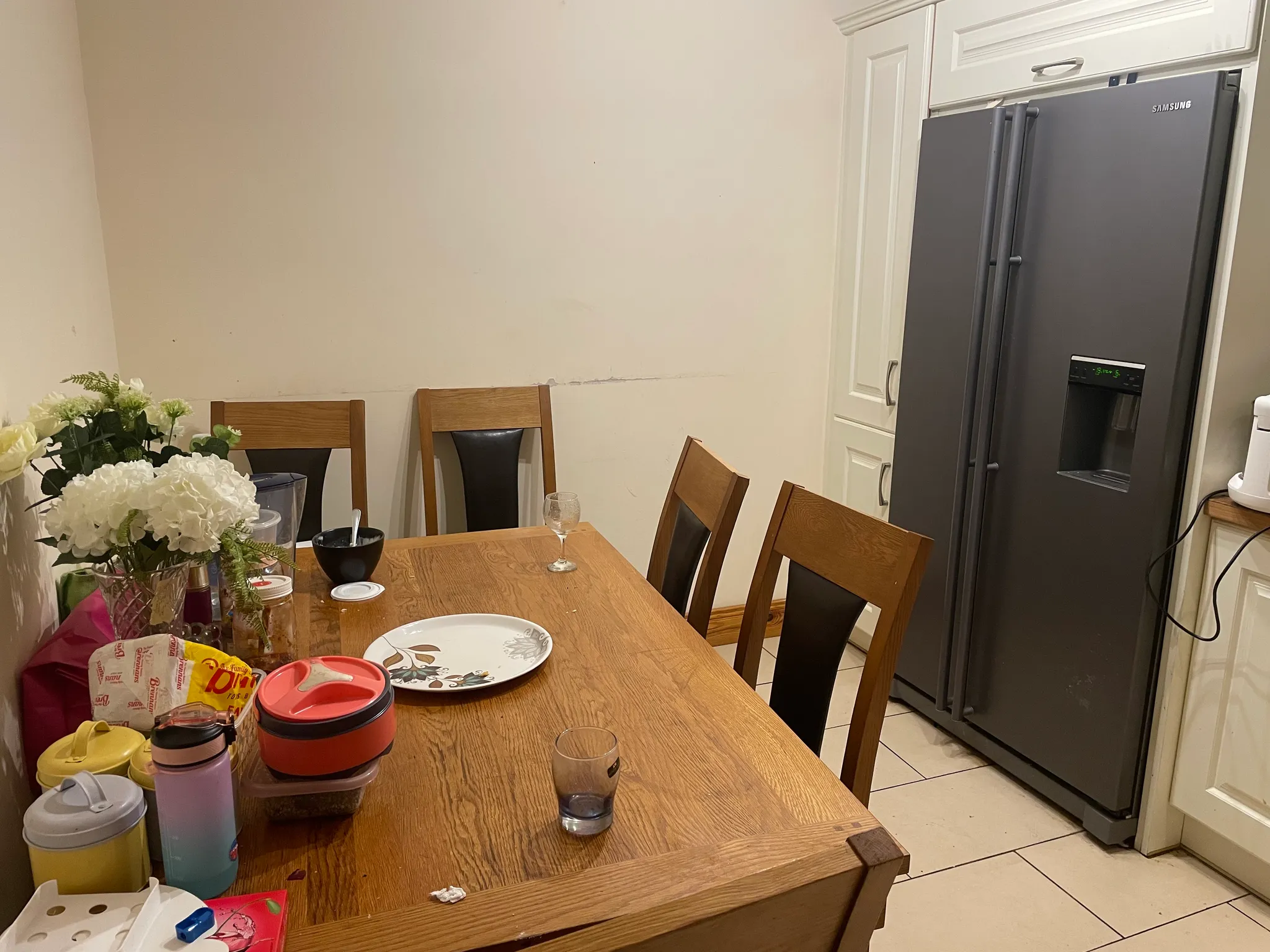 Dining area with a wooden table and chairs, assorted dishes and containers on the table, a vase of flowers at left, and a stainless refrigerator and cabinets at right.