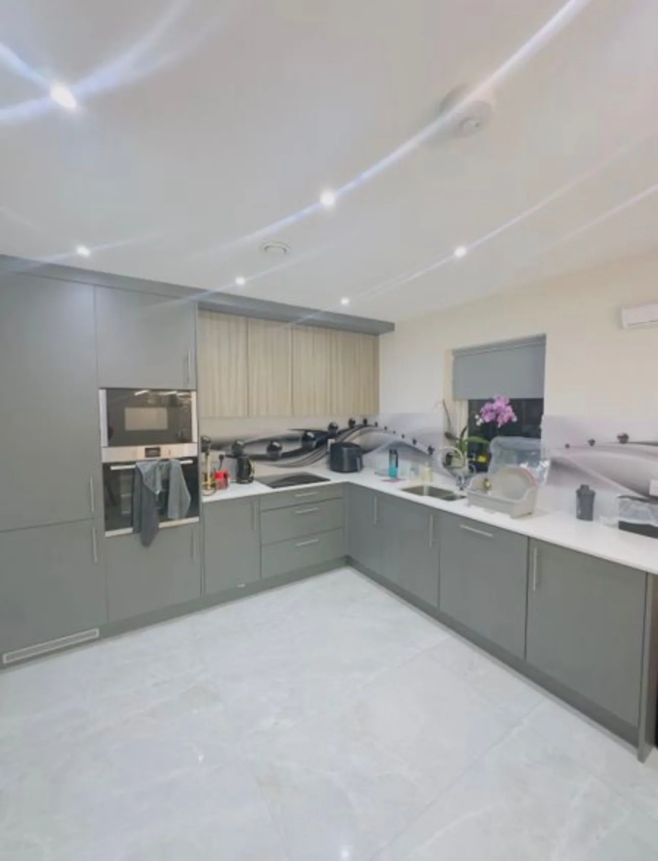 Kitchen with grey cabinets, L-shaped white countertop, built-in oven, sink, small window and appliances on the counter