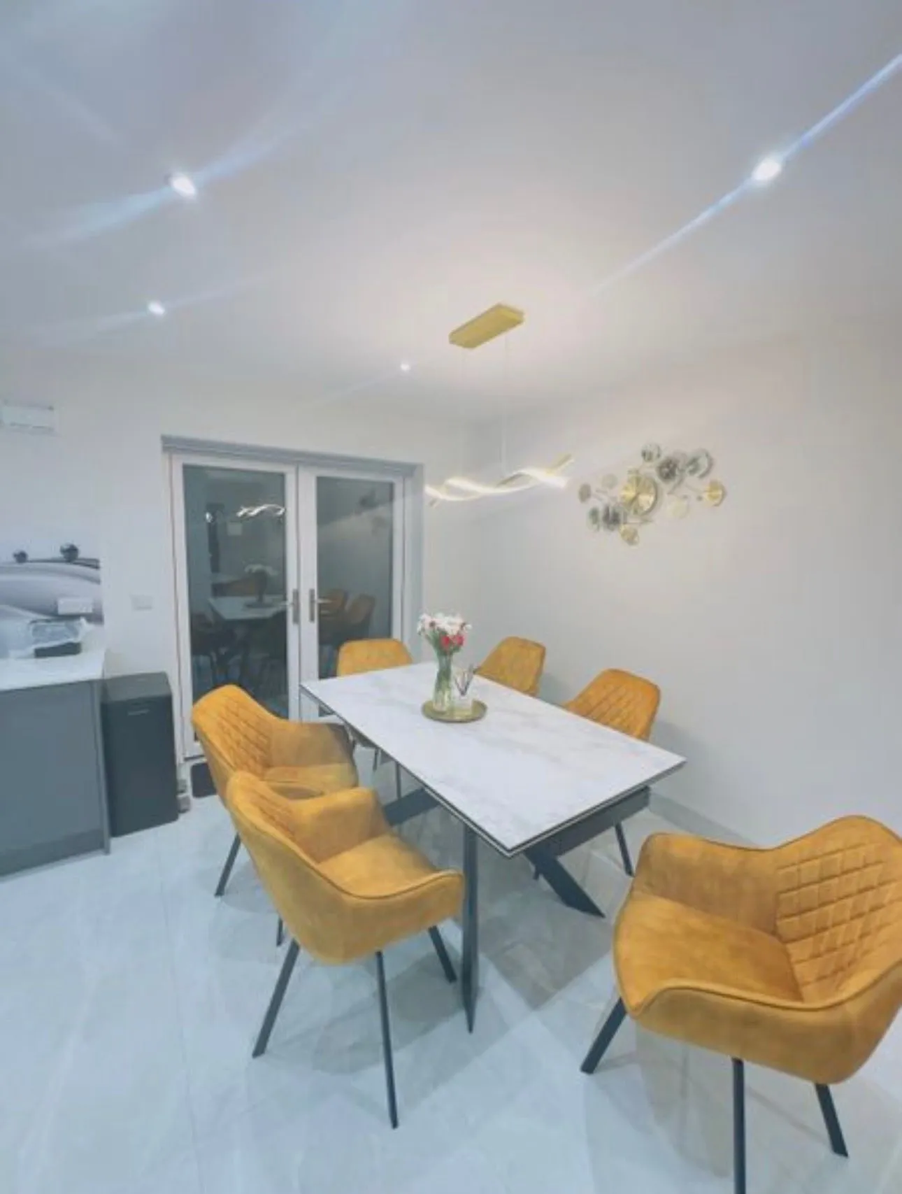 Dining room with a rectangular table, six mustard-yellow upholstered chairs, a vase of flowers on the table, pendant light fixture, and sliding glass doors.