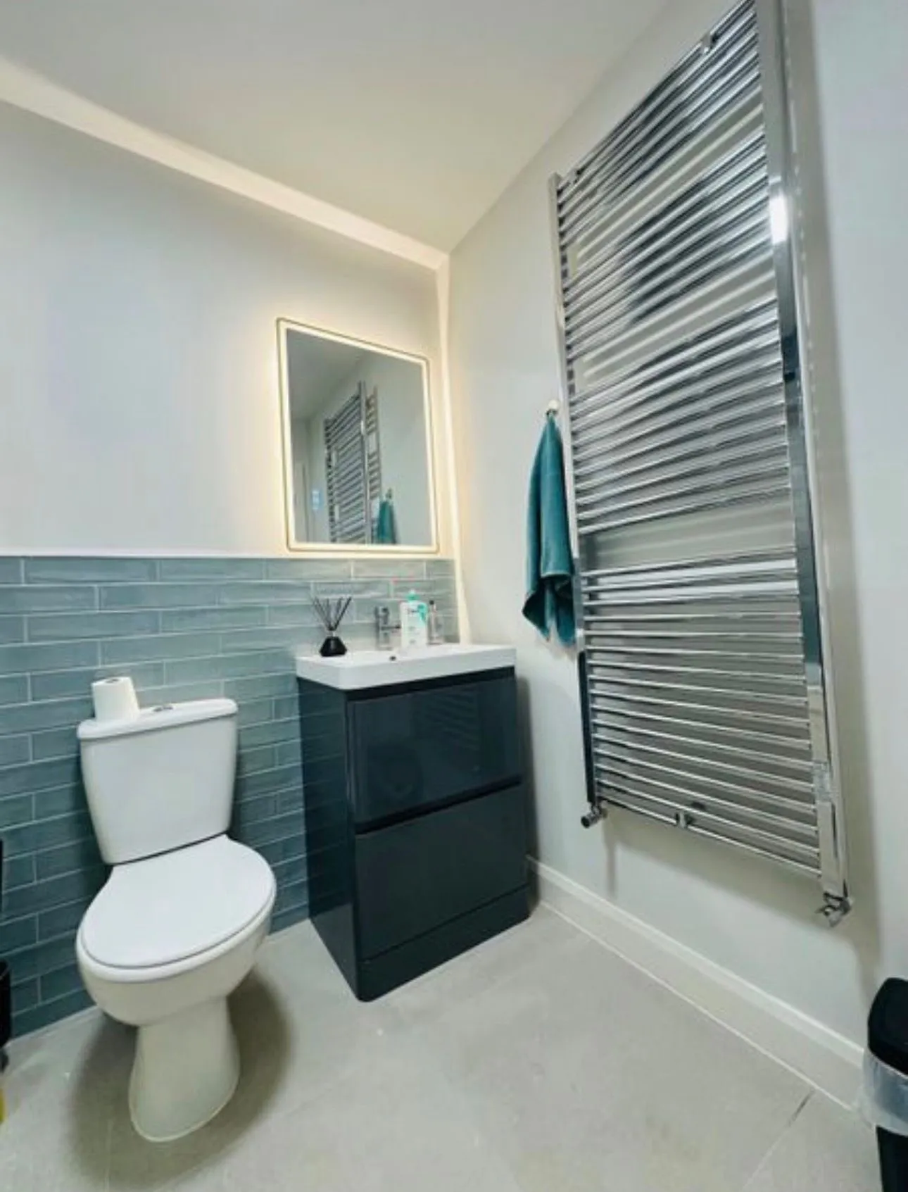 Bathroom with a toilet, dark vanity with sink and illuminated mirror, wall-mounted chrome towel radiator and a teal towel