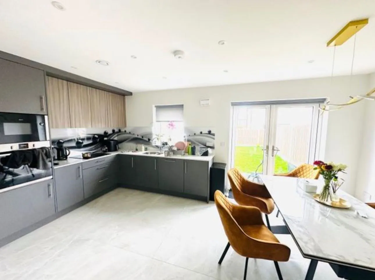 Kitchen with L-shaped gray cabinets, built-in oven, sink under a small window, marble-top dining table and three brown upholstered chairs next to glass patio doors.
