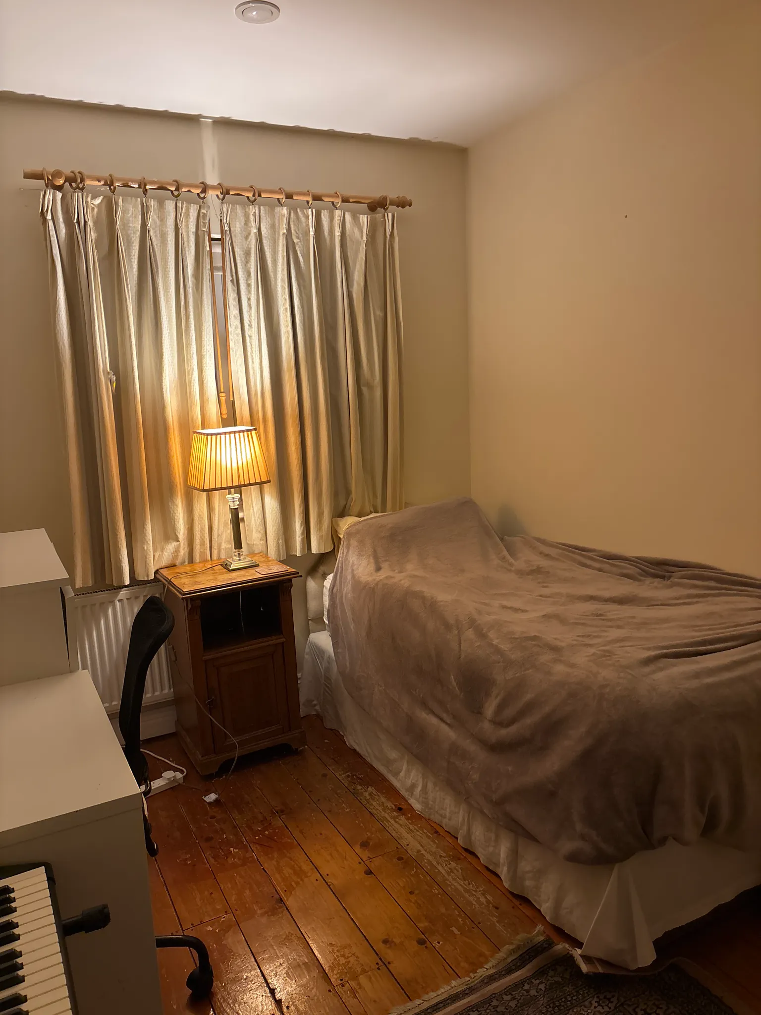 Bedroom with a single bed covered by a blanket, closed curtains, a bedside table with a lamp, and wooden floorboards.