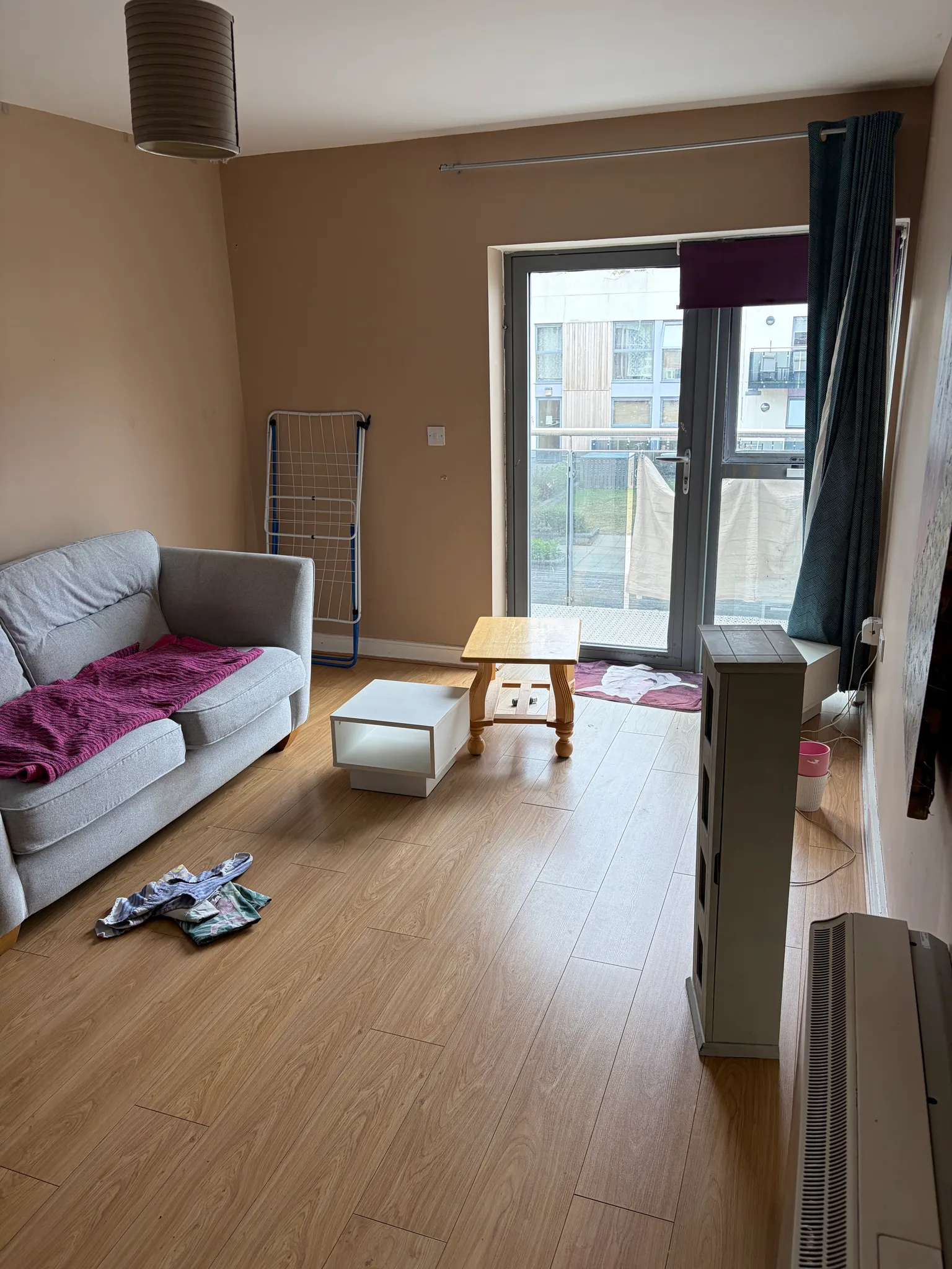 Living room with a sofa, small coffee tables, floor-to-ceiling glass door to a balcony, curtains and a freestanding drying rack.