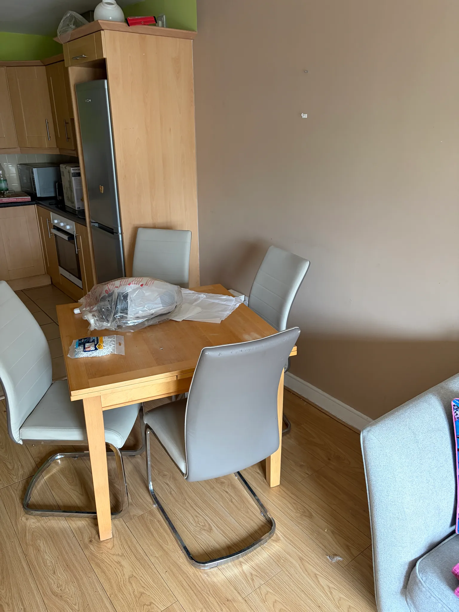 Dining area with a wooden table, four grey dining chairs and a plastic-wrapped item on the table; kitchen cabinets and a refrigerator visible to the left and a plain beige wall to the right.
